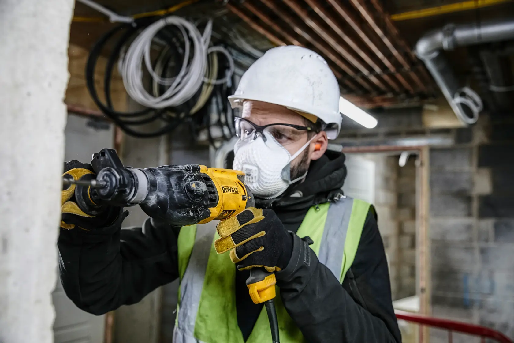 Una persona con casco de seguridad, guantes y chaleco reflectante está usando un taladro DEWALT para trabajar en una pared de hormigón en un entorno de construcción. El rostro está difuminado por privacidad.