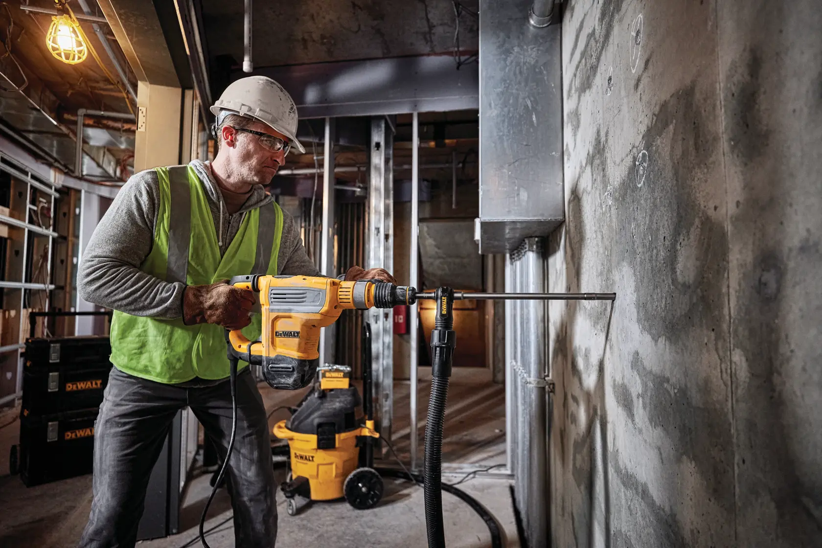 Una persona con equipo de seguridad y casco utiliza un martillo rotativo DEWALT D25614K sobre una pared de concreto en un sitio de construcción. Se pueden ver equipos y herramientas DEWALT en el fondo.