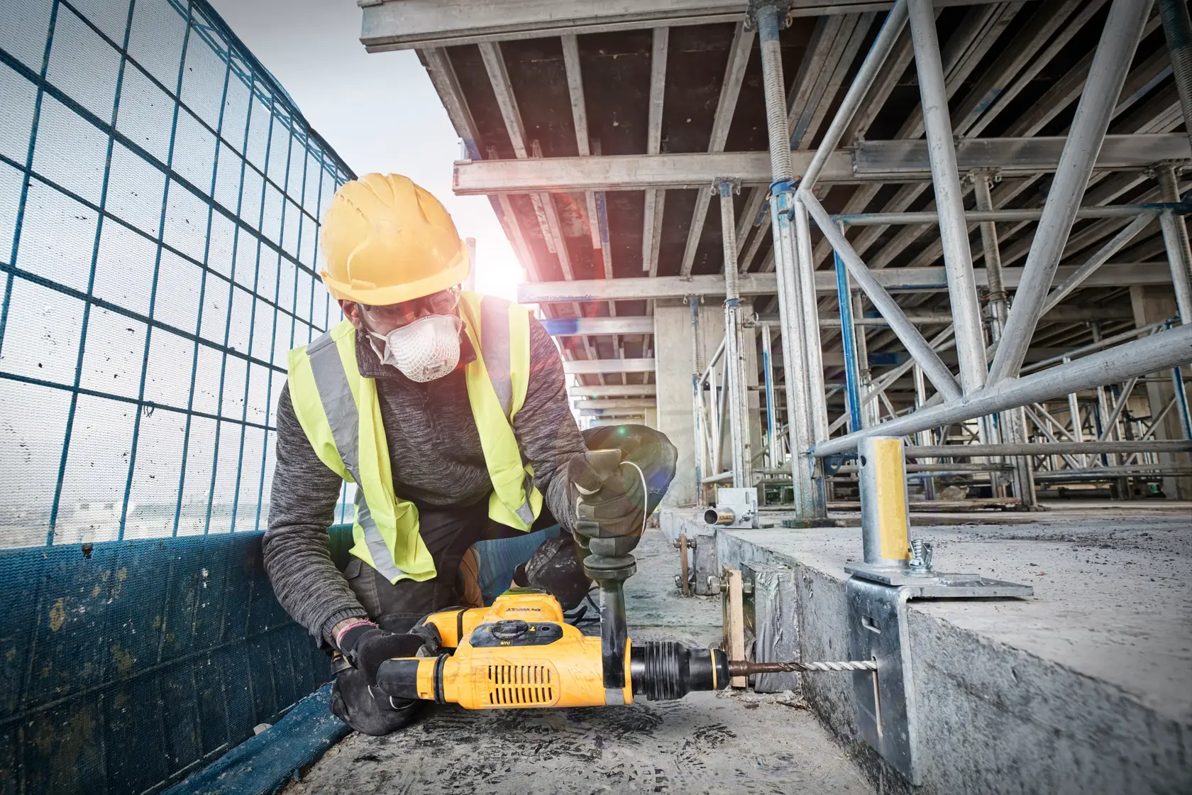 Un trabajador de la construcción con casco amarillo y chaleco reflectante está utilizando un martillo perforador amarillo DEWALT para perforar una superficie de hormigón en una obra.