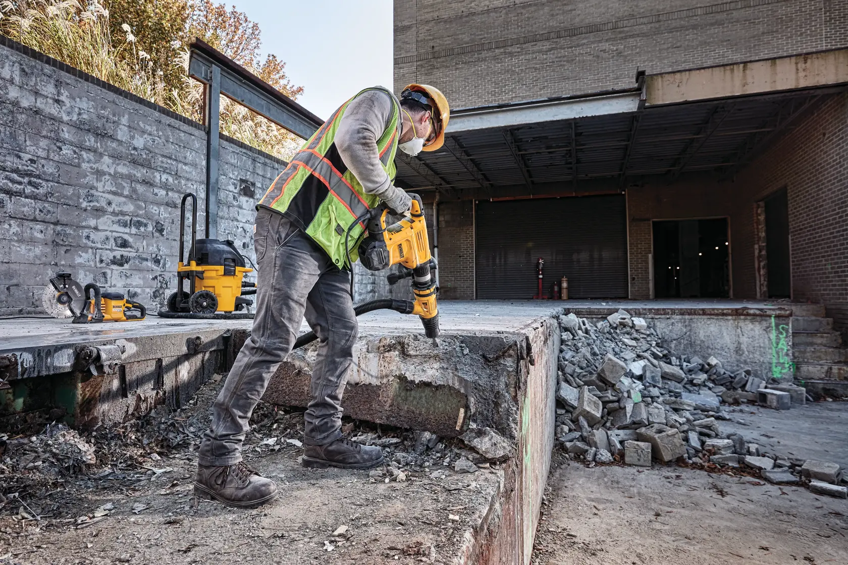 Un trabajador de construcción con equipo de seguridad utiliza un martillo de demolición DEWALT en una estructura de concreto en una obra. Se puede ver concreto desmoronado y escombros alrededor del área.