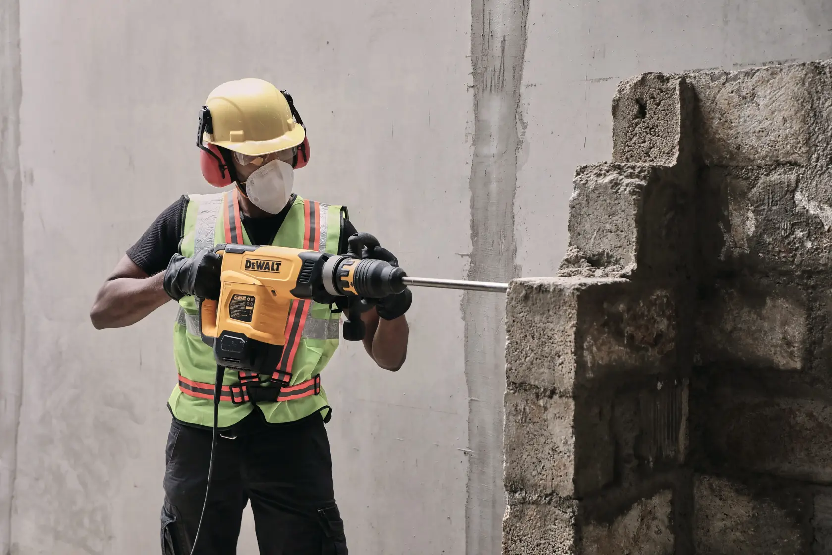 Un trabajador de la construcción con casco de seguridad amarillo, protección auditiva, mascarilla y chaleco reflectante utiliza un martillo demoledor DEWALT para romper una pared de concreto.