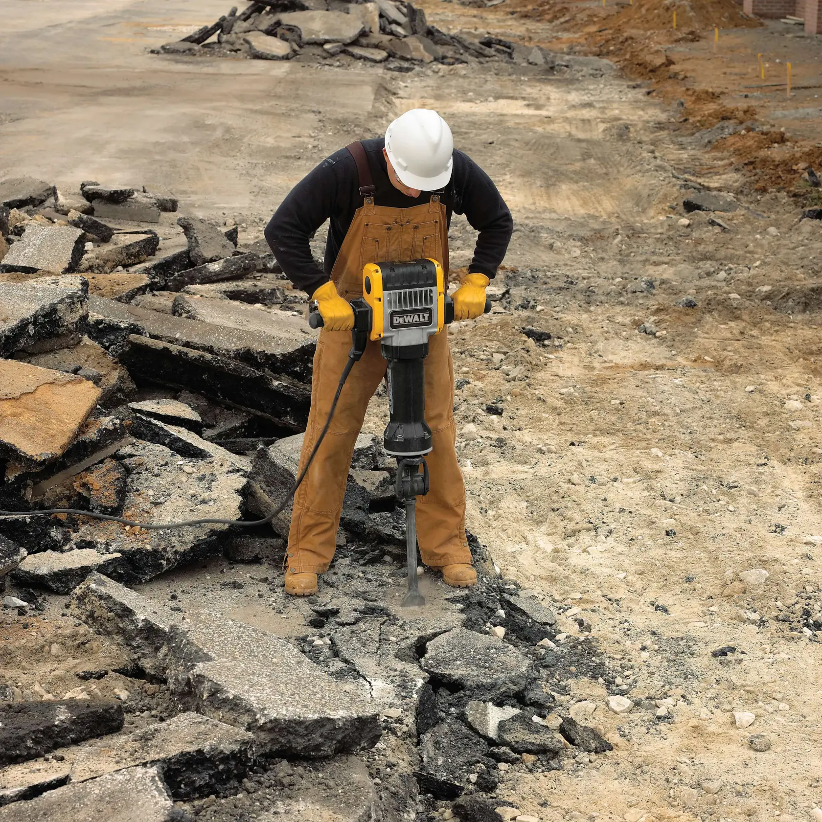 Um trabalhador usando capacete e luvas utiliza um martelete DEWALT para quebrar concreto ou asfalto em um canteiro de obras.