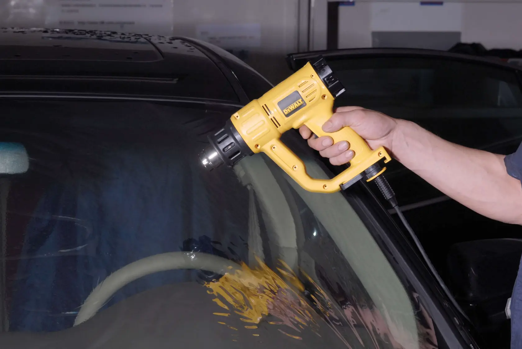 Una persona utiliza una pistola de aire caliente DEWALT (modelo D26411) para aplicar calor en el parabrisas de un coche.