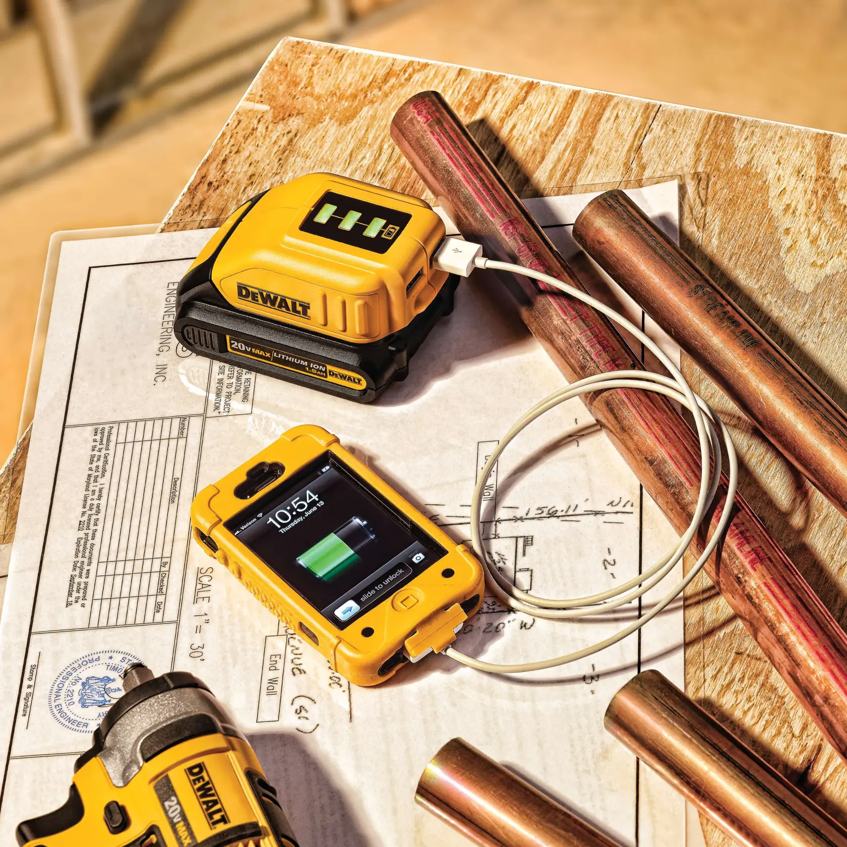 Un cargador de batería de ion de litio DEWALT 20V está cargando un smartphone mediante un cable USB. El cargador y el teléfono están sobre planos de ingeniería y una superficie de madera, rodeados de tubos de cobre y otras herramientas DEWALT.
