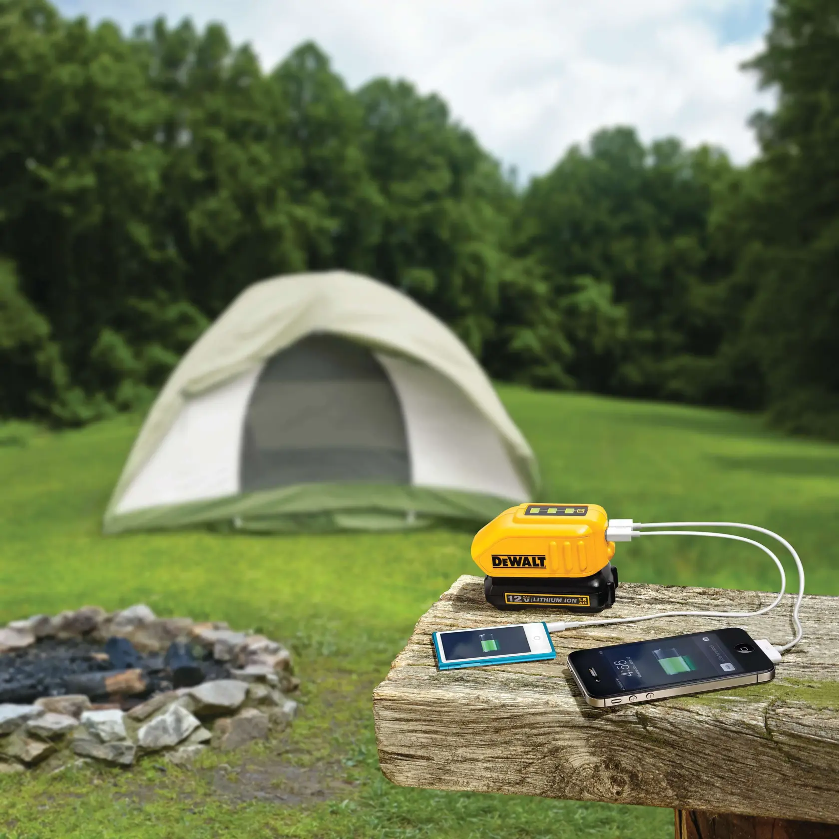 Un dispositivo de carga portátil DEWALT está alimentando dos dispositivos electrónicos sobre una mesa de madera al aire libre, con una tienda de campaña y una fogata al fondo rodeados de césped y árboles.