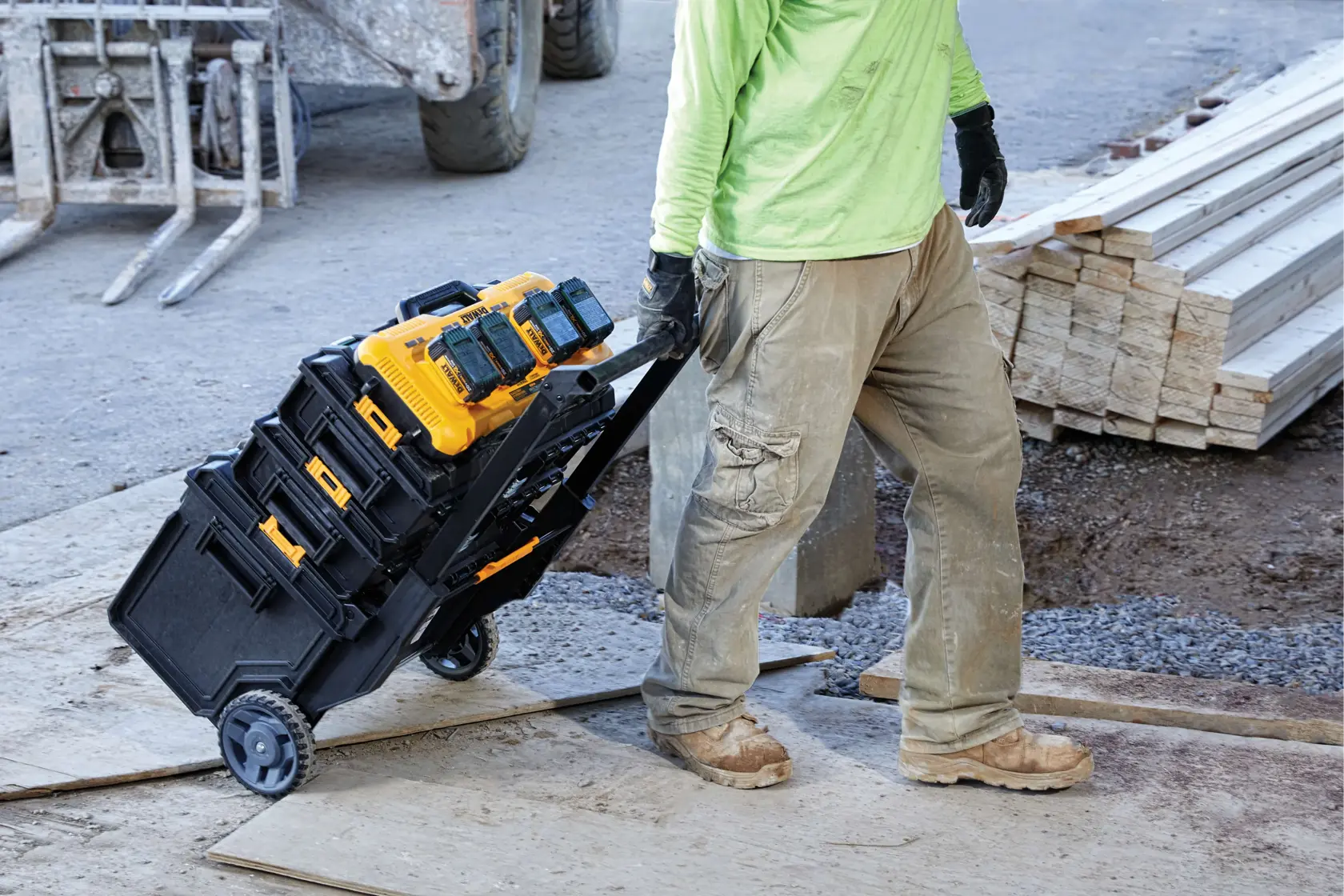 Un trabajador con ropa de trabajo tira de un carro de herramientas DEWALT negro y amarillo, cargado con cargadores de baterías inalámbricas y cajas de herramientas, a través de una obra en construcción, con madera y materiales de construcción al fondo.