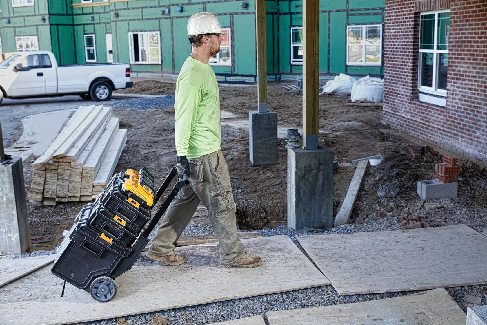 Un trabajador de la construcción con casco y guantes tira de un carro de herramientas con varias cajas de herramientas DEWALT en una obra. Al fondo se aprecia un edificio en construcción y materiales de construcción alrededor.