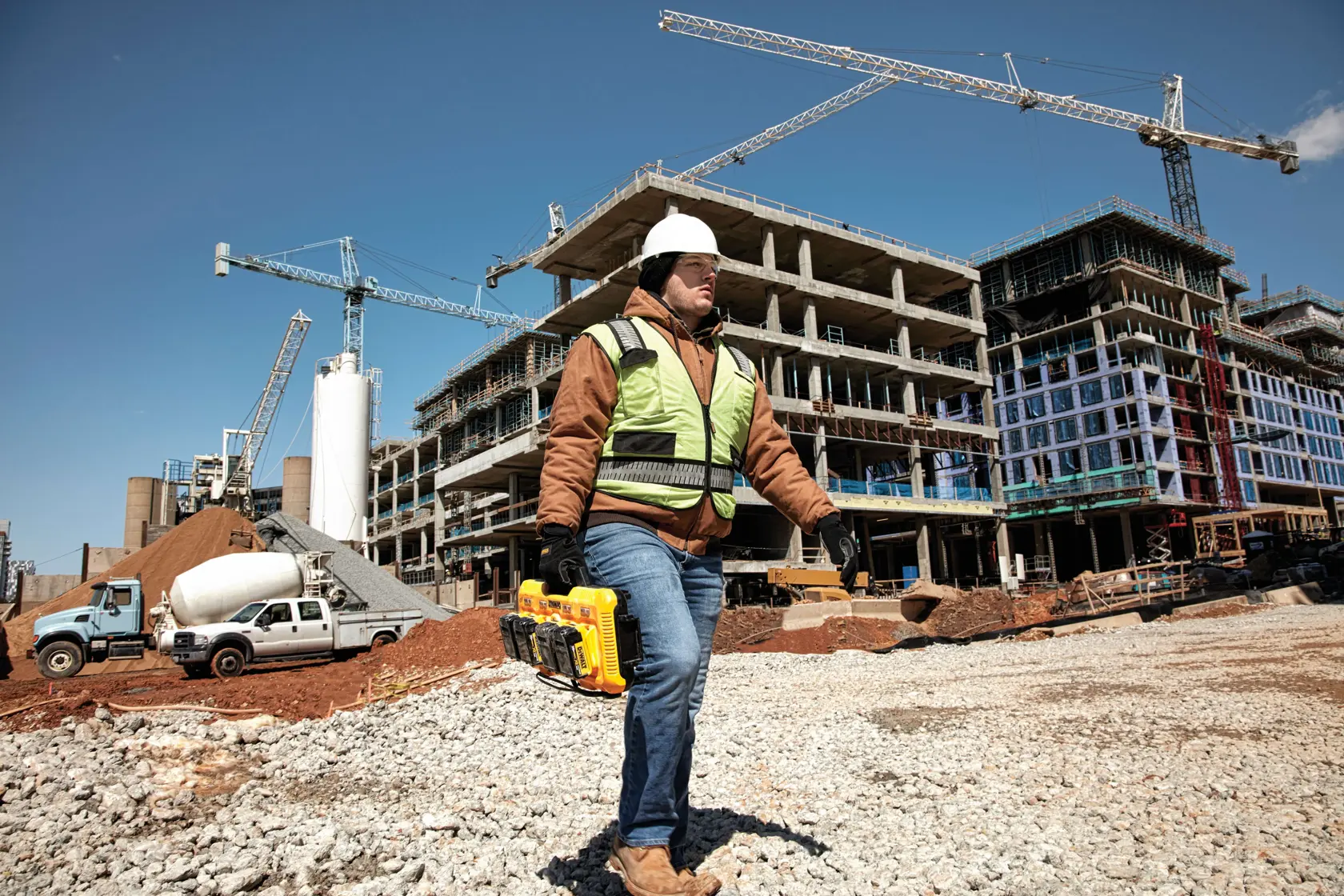 Un trabajador de construcción con chaleco de seguridad y casco lleva un cargador de batería DEWALT DCB104 en una gran obra con edificios sin terminar, grúas y vehículos de construcción al fondo.