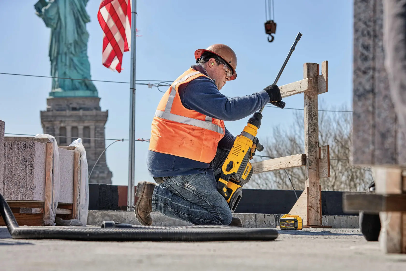 Un trabajador de la construcción con chaleco de seguridad naranja y casco utiliza una herramienta eléctrica amarilla DEWALT para perforar en un sitio de obra. El trabajador está arrodillado cerca de estructuras de madera, con la batería DEWALT DCB612 visible junto a él. Al fondo se ven la Estatua de la Libertad y una bandera estadounidense.