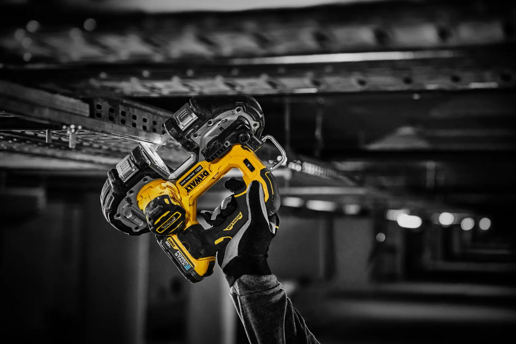 Una persona con guante sostiene una herramienta eléctrica inalámbrica amarilla DEWALT bajo una estructura metálica, posiblemente demostrando su uso en un entorno de construcción o industrial.