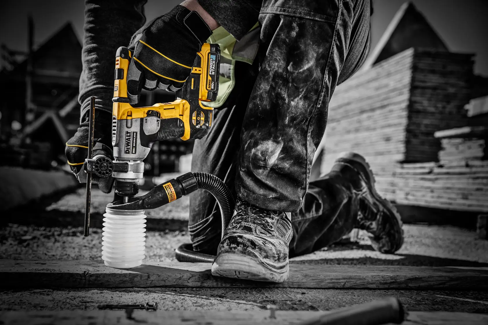 Un trabajador con guantes y botas de trabajo utiliza una herramienta eléctrica inalámbrica amarilla y negra DEWALT con una batería DCBP034 en un sitio de construcción. La herramienta se está utilizando para perforar concreto o piedra y tiene un accesorio de extracción de polvo conectado.