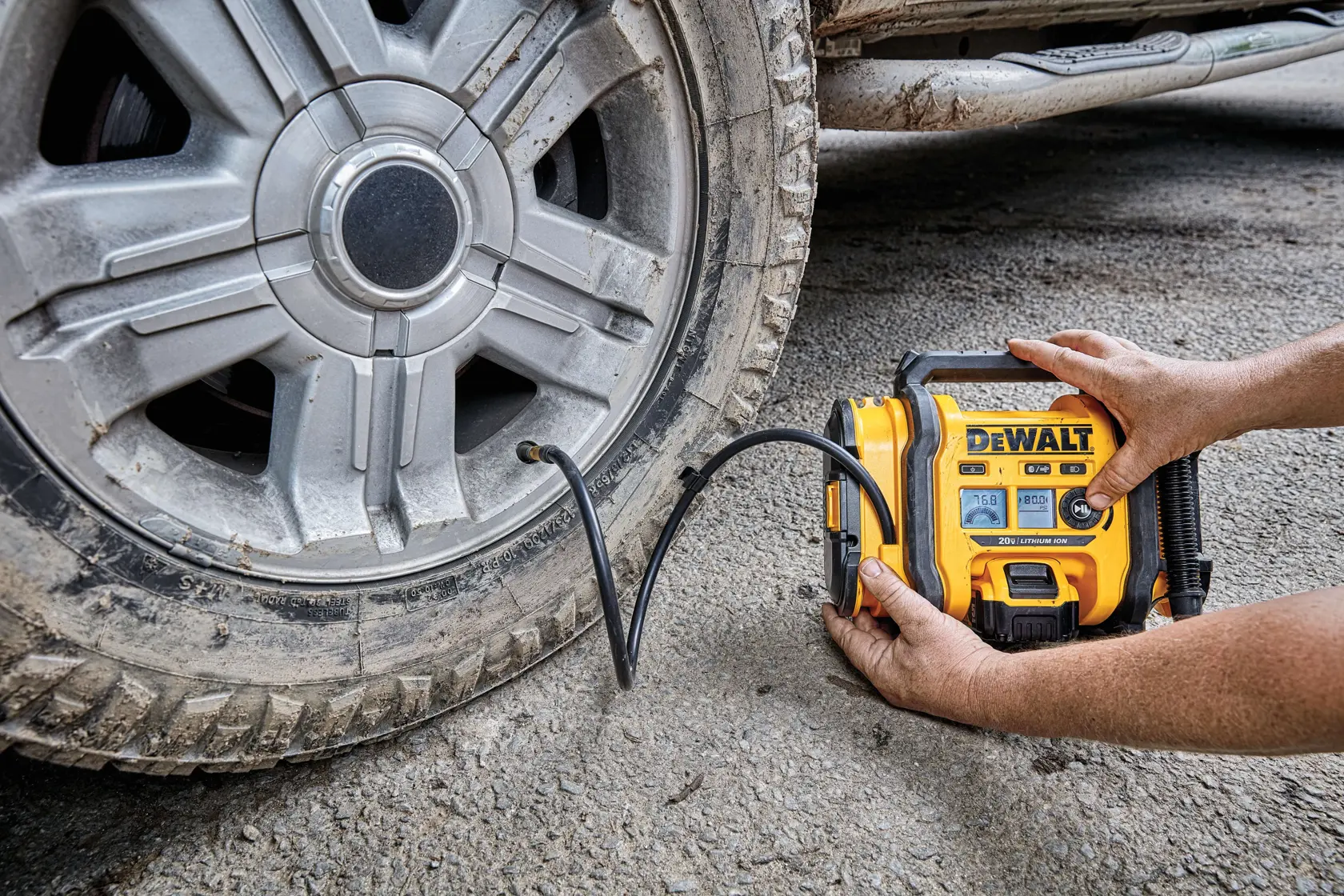 Una persona está utilizando un compresor de aire portátil amarillo DEWALT para inflar una llanta de coche sobre una superficie exterior irregular. Se puede ver la pantalla digital y la manguera del compresor.