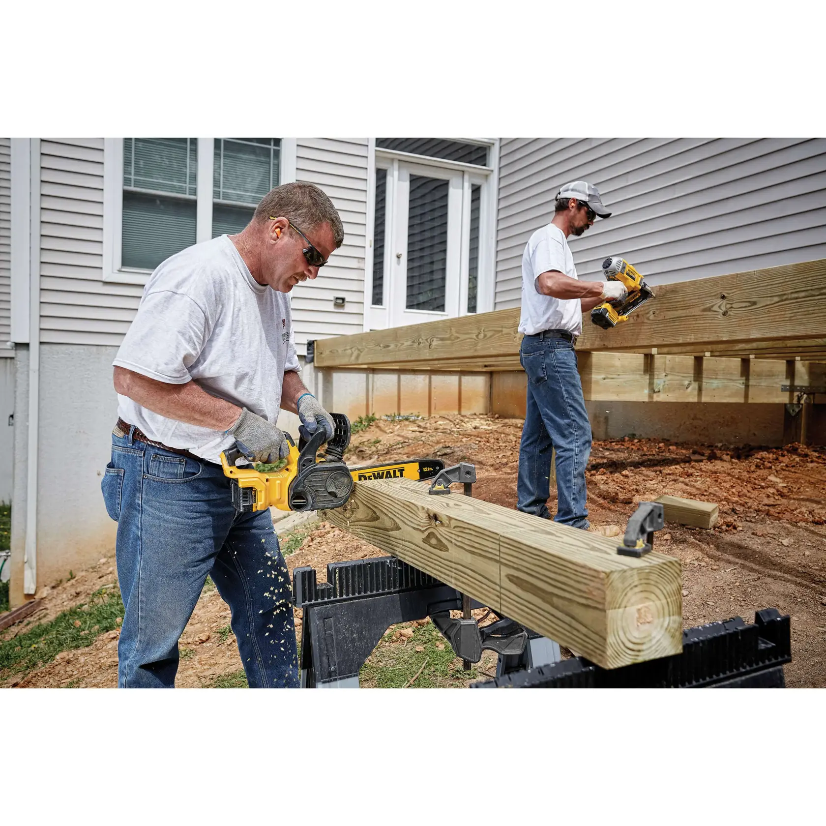 Una persona con guantes y camiseta gris usa una motosierra inalámbrica DEWALT (modelo DCCS620B-B3) para cortar una gran viga de madera al aire libre, con astillas de madera volando. Se ve una casa y una terraza sin terminar en el fondo.