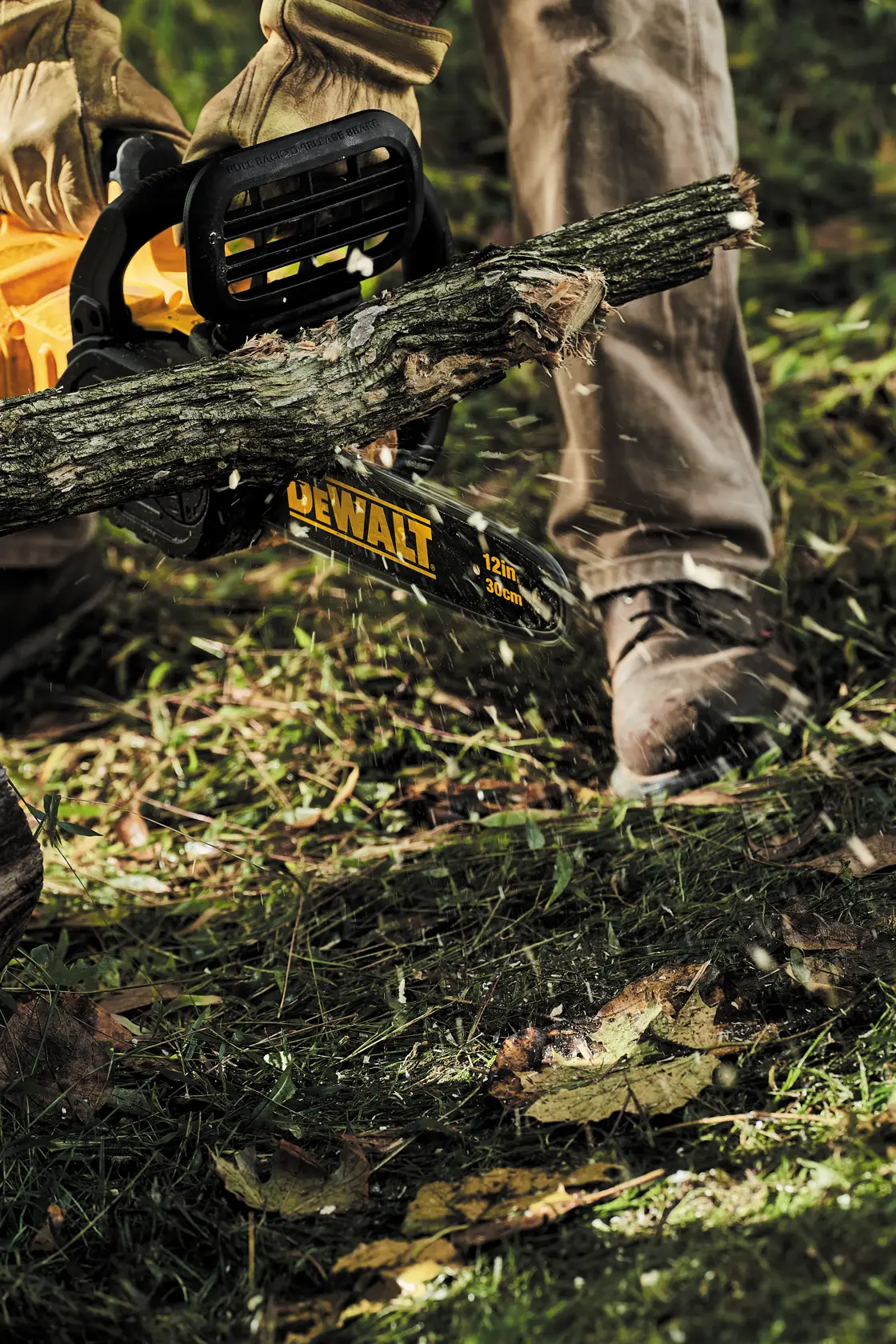 Una persona con guantes y ropa protectora está usando una motosierra DEWALT de 12 pulgadas para cortar una rama de árbol al aire libre, con astillas de madera volando y césped debajo.