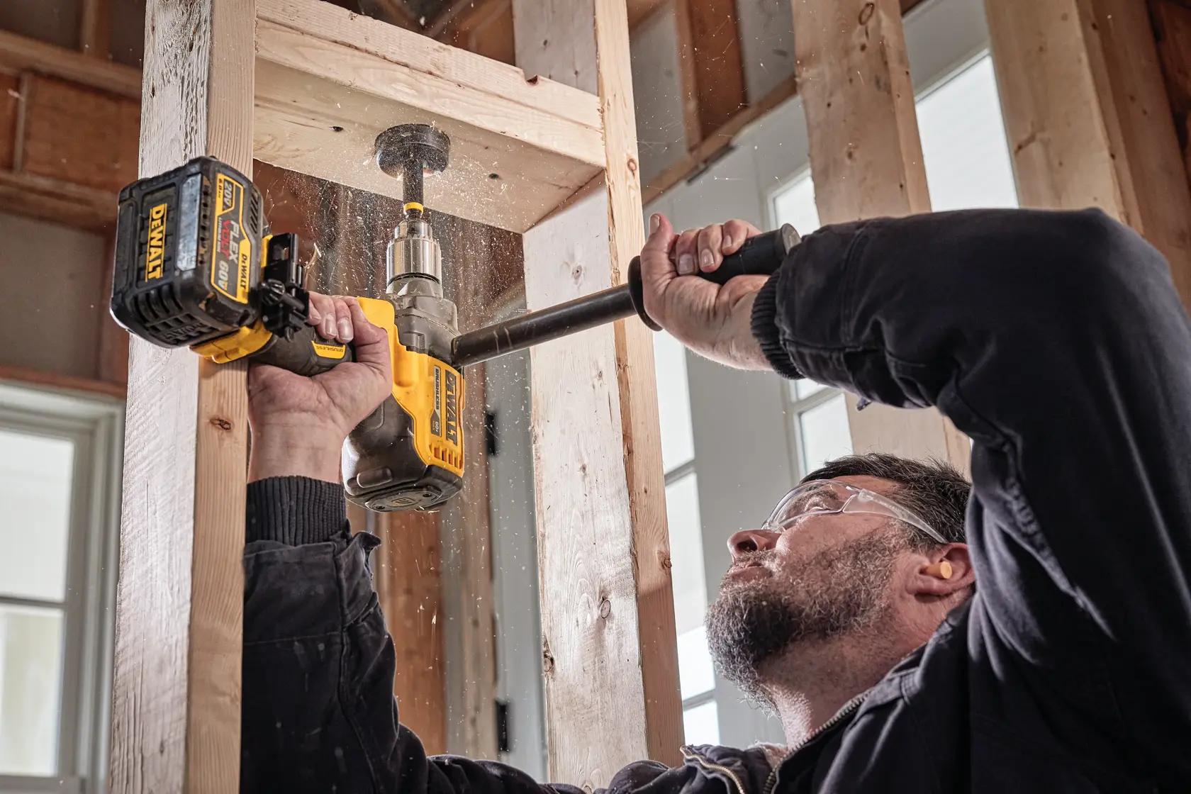 Persona usando un taladro inalámbrico DEWALT para perforar un agujero en vigas de madera dentro de un edificio en construcción. El taladro se sostiene con ambas manos y se ven partículas de aserrín.