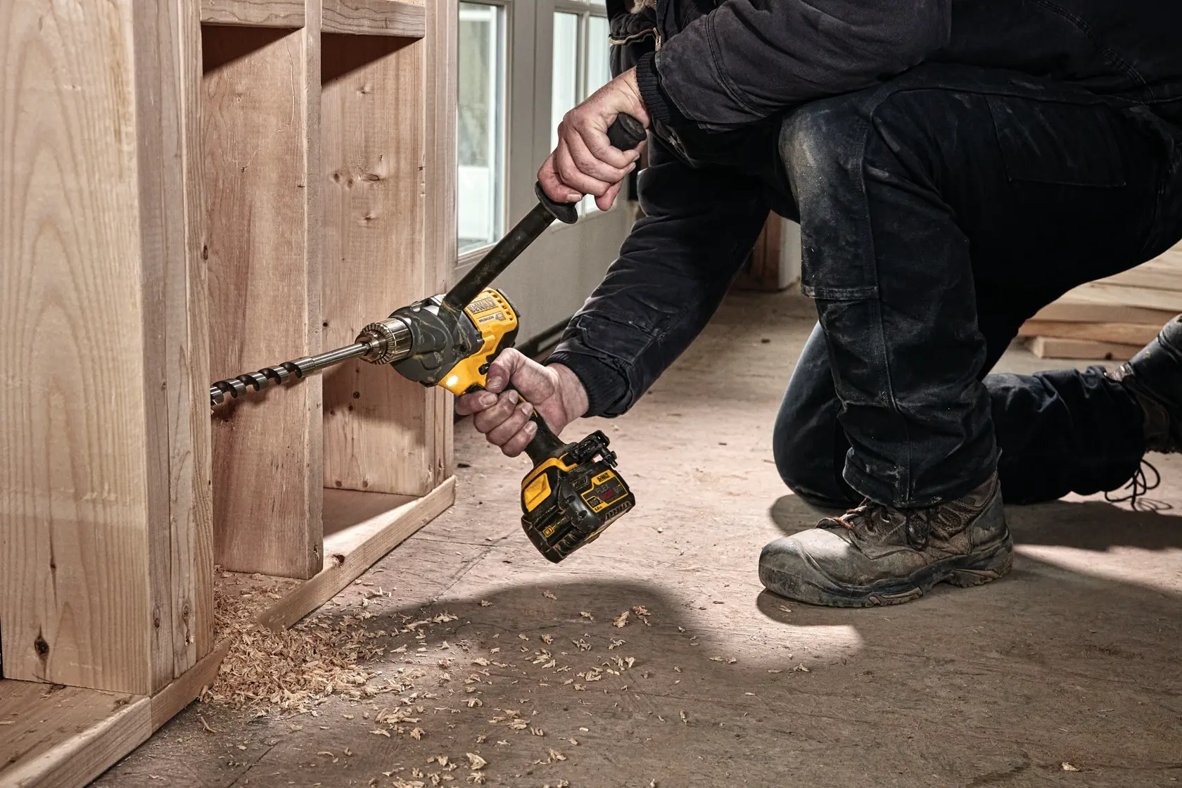 Una persona está arrodillada en el suelo usando un taladro inalámbrico DEWALT con una broca larga para perforar agujeros en un gabinete o estructura de madera. Virutas de madera están esparcidas en el suelo.
