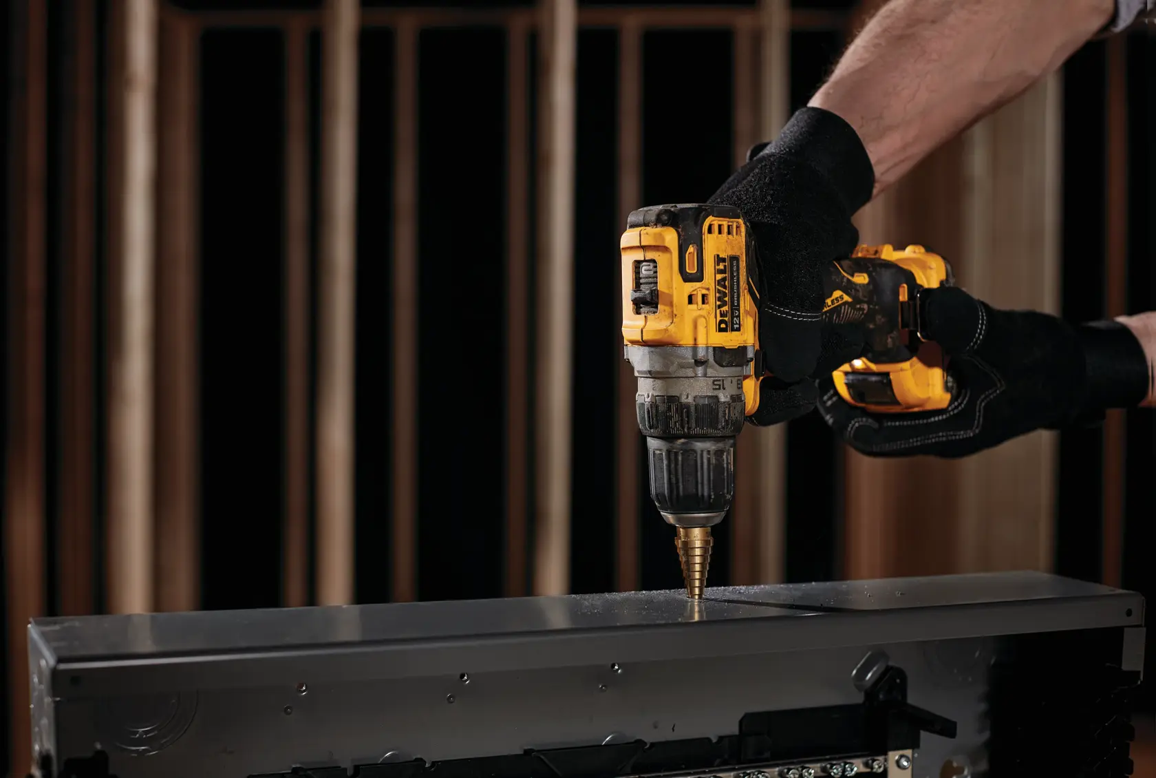 Una persona con guantes negros utiliza un taladro inalámbrico amarillo DEWALT para perforar una pieza de metal.
