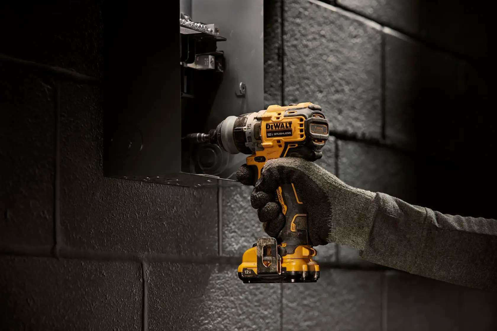 Una persona con guantes de trabajo utiliza un taladro inalámbrico amarillo y negro DEWALT para trabajar en un panel eléctrico montado en una pared de ladrillo.