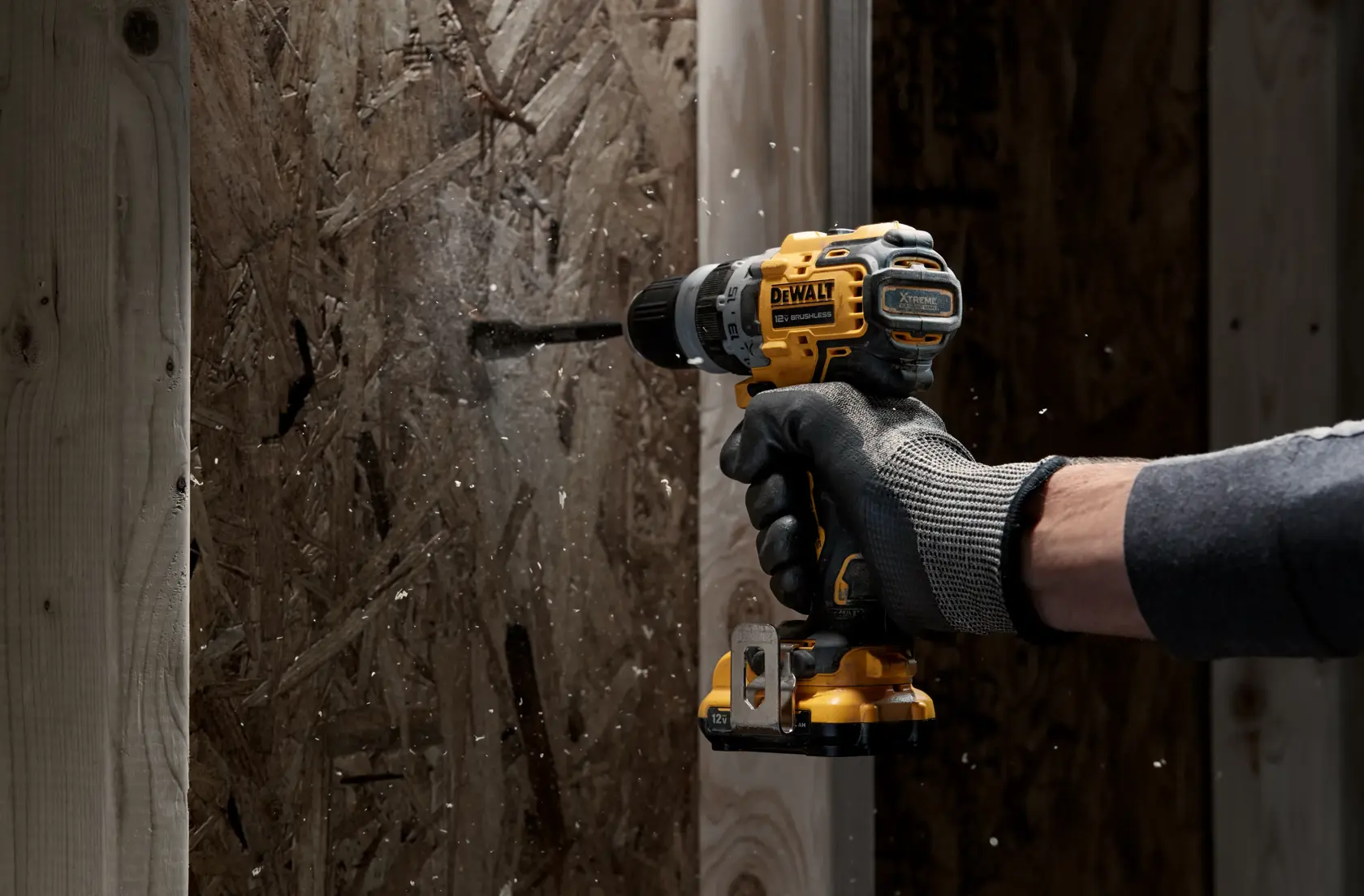 Una persona con guantes protectores utiliza un taladro inalámbrico DEWALT para perforar un panel de madera, generando astillas y polvo de madera.