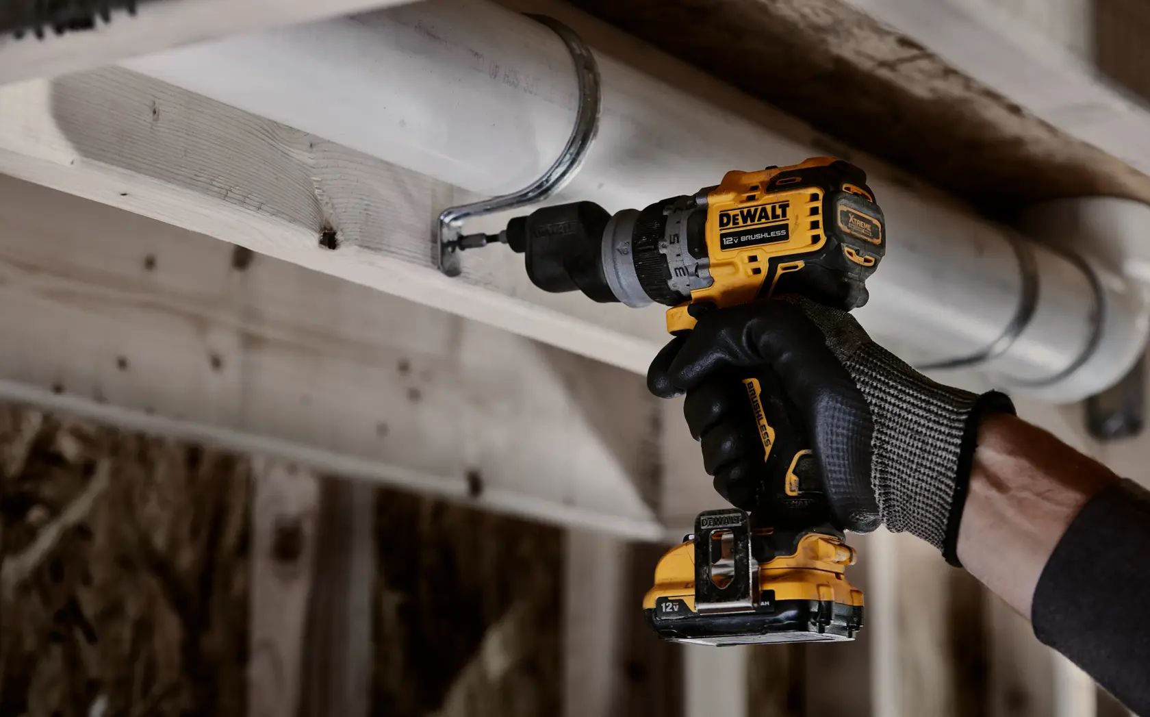 Una persona con guantes de trabajo utiliza un taladro inalámbrico DEWALT para asegurar un soporte metálico alrededor de una tubería blanca fijada a una viga de madera.