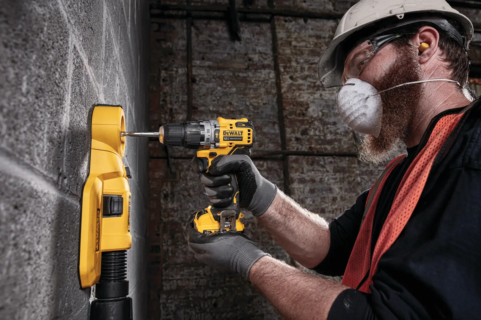 Una persona con equipo de protección opera un taladro inalámbrico DEWALT mientras perfora una pared de concreto. Un extractor de polvo amarillo está acoplado a la pared para recolectar el polvo durante el proceso.