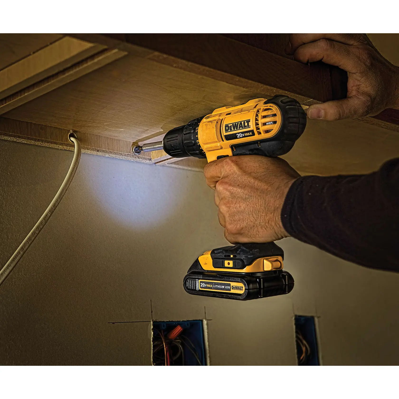 Una persona está usando un taladro inalámbrico DEWALT de 20V (modelo DCD771C2) para perforar o atornillar en la parte inferior de un gabinete de madera. El taladro es amarillo y negro, y el área está iluminada por la luz LED incorporada.