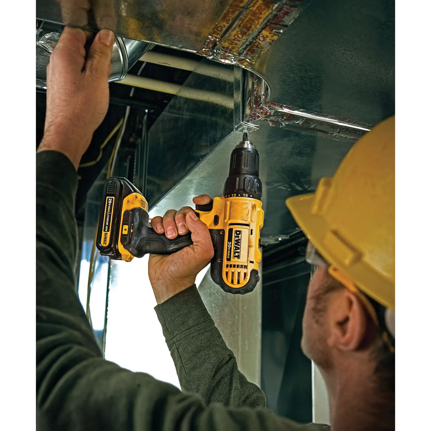 Una persona con casco de seguridad amarillo utiliza un taladro inalámbrico DEWALT para fijar o perforar un conducto metálico, sosteniendo el taladro con una mano y estabilizando el metal con la otra.