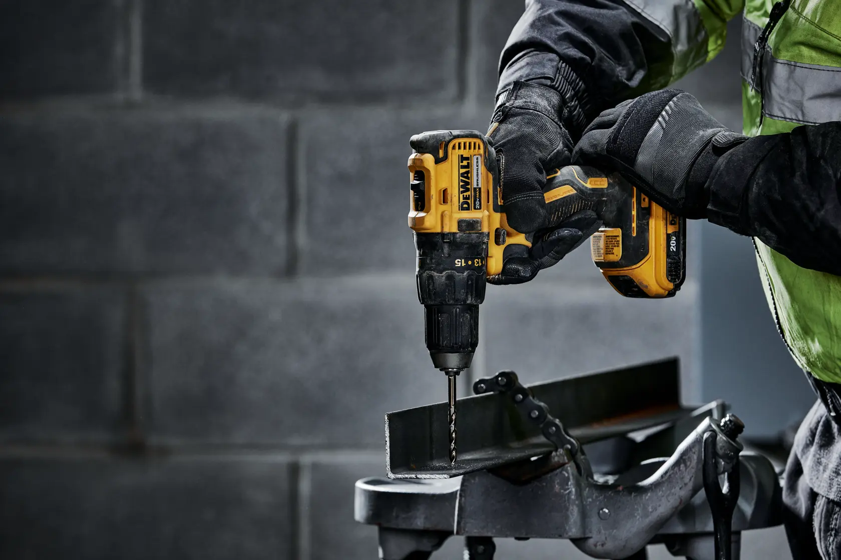 Una persona con guantes de protección utiliza un taladro inalámbrico amarillo DEWALT para perforar una pieza de metal sujeta en un tornillo de banco de metal.