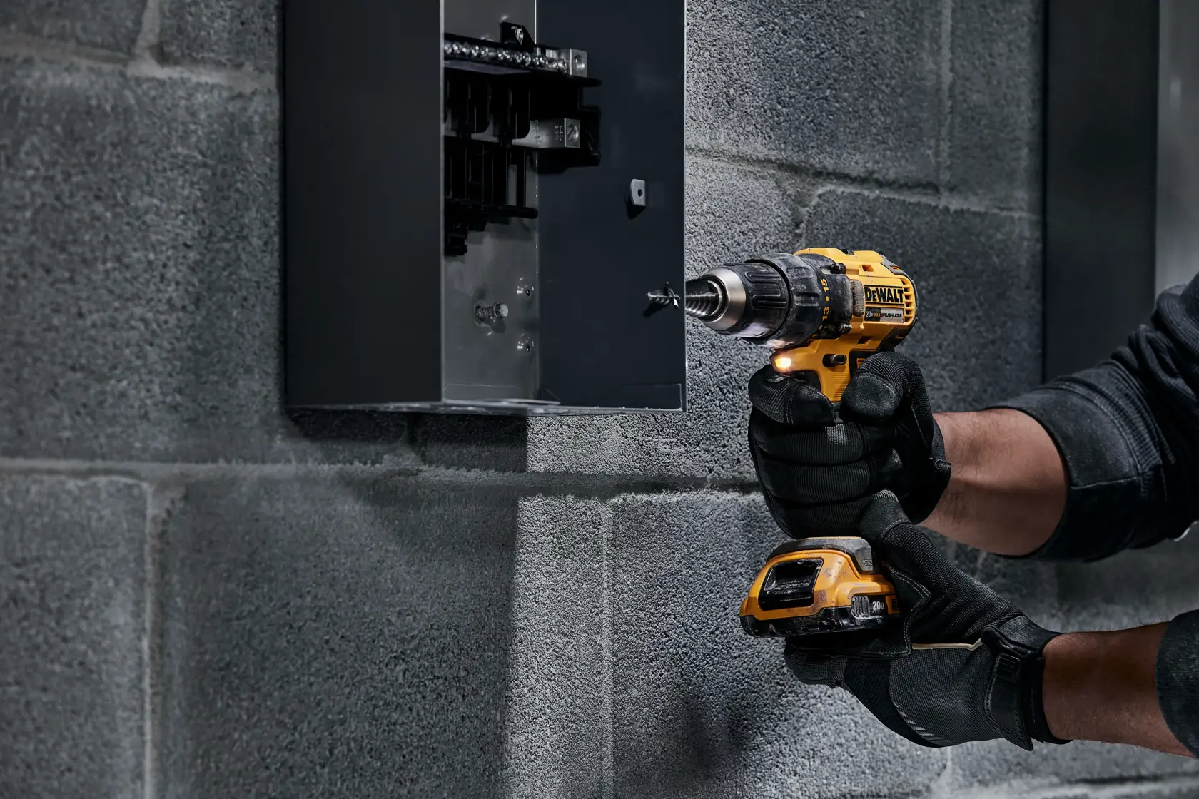 Una persona con guantes negros está usando un taladro inalámbrico amarillo DEWALT para trabajar en un panel eléctrico gris montado en una pared de concreto.