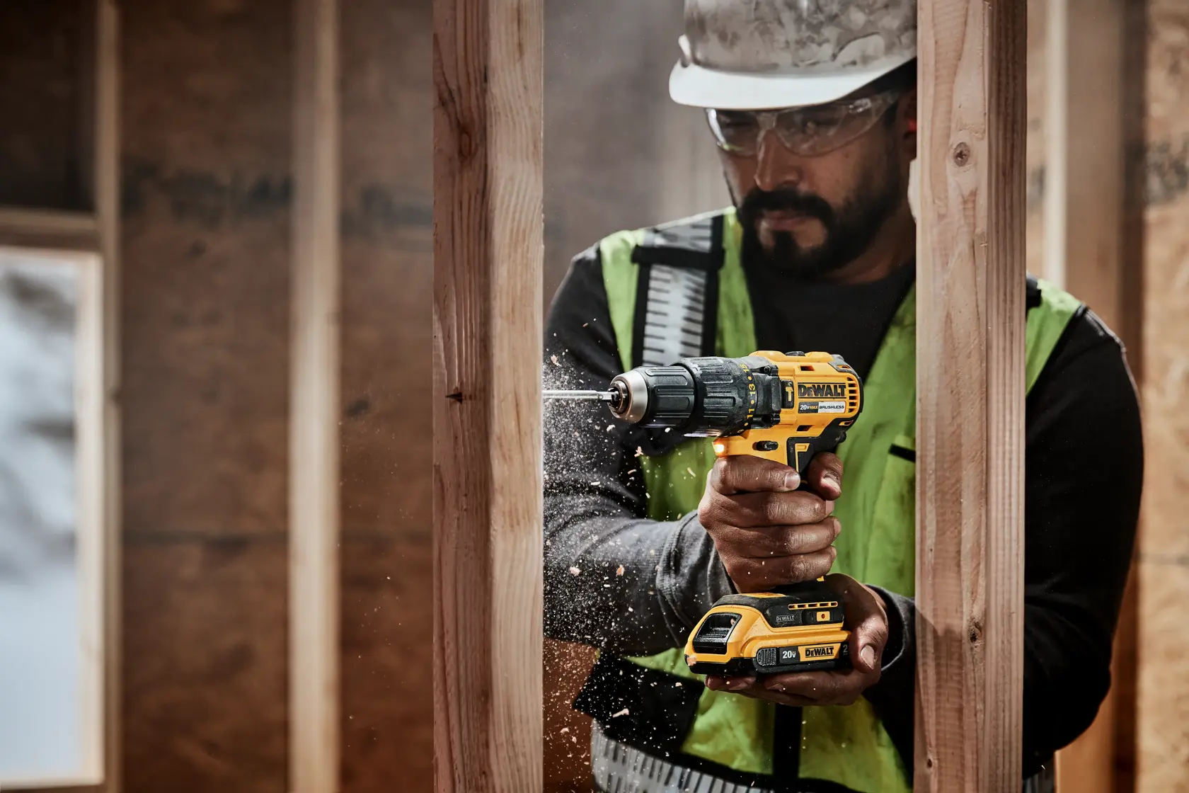 Una persona con chaleco de seguridad y casco utiliza un taladro inalámbrico DEWALT para perforar una viga de madera en un sitio de construcción. Se pueden ver virutas de madera alrededor de la broca.