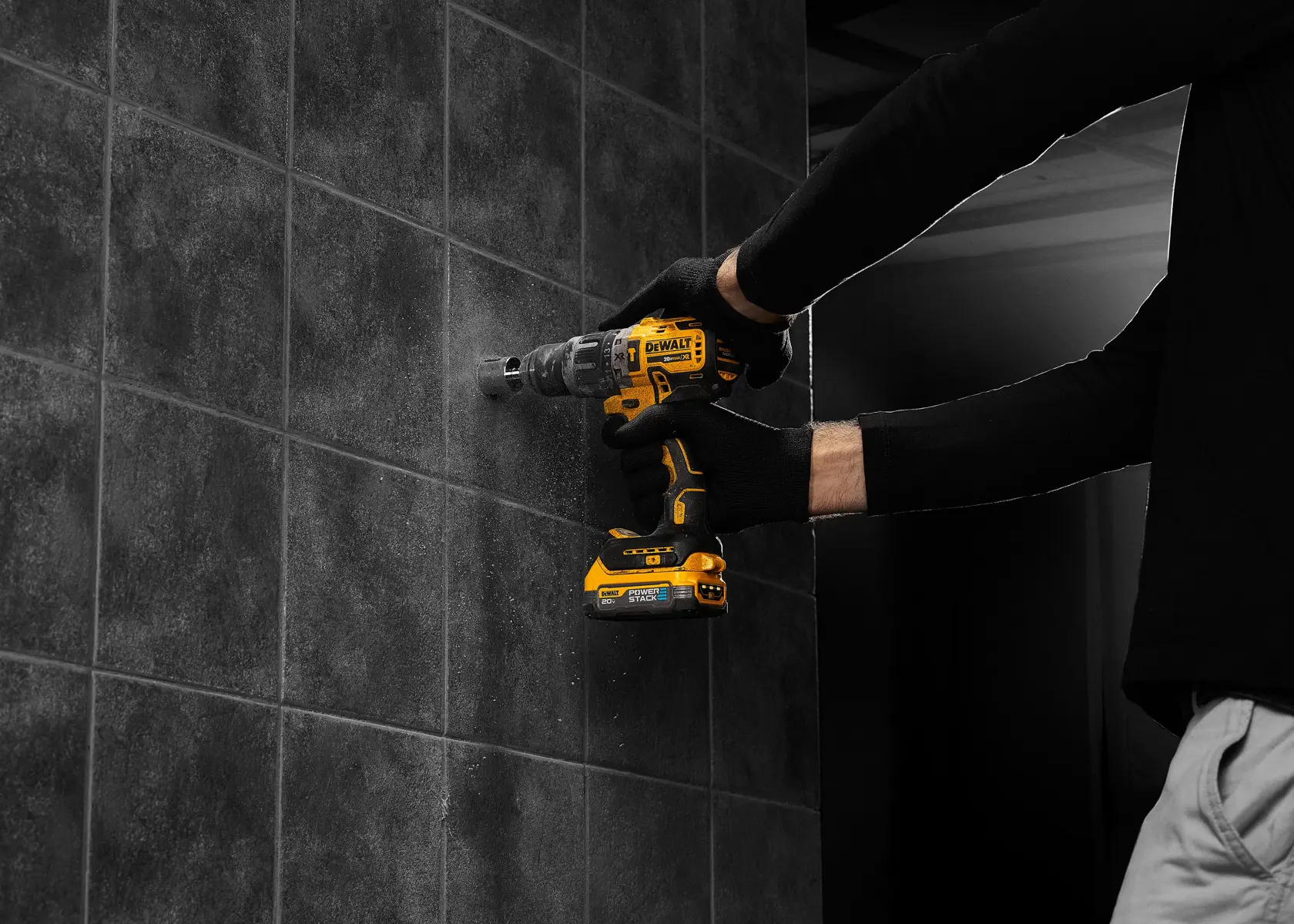 Una persona con guantes negros utiliza un taladro inalámbrico DEWALT para hacer un agujero en una pared de baldosas oscuras.
