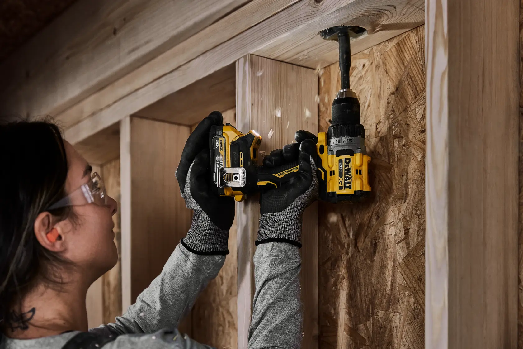 Una persona con guantes de trabajo está usando un taladro inalámbrico DEWALT para hacer un agujero en un marco o pared de madera.
