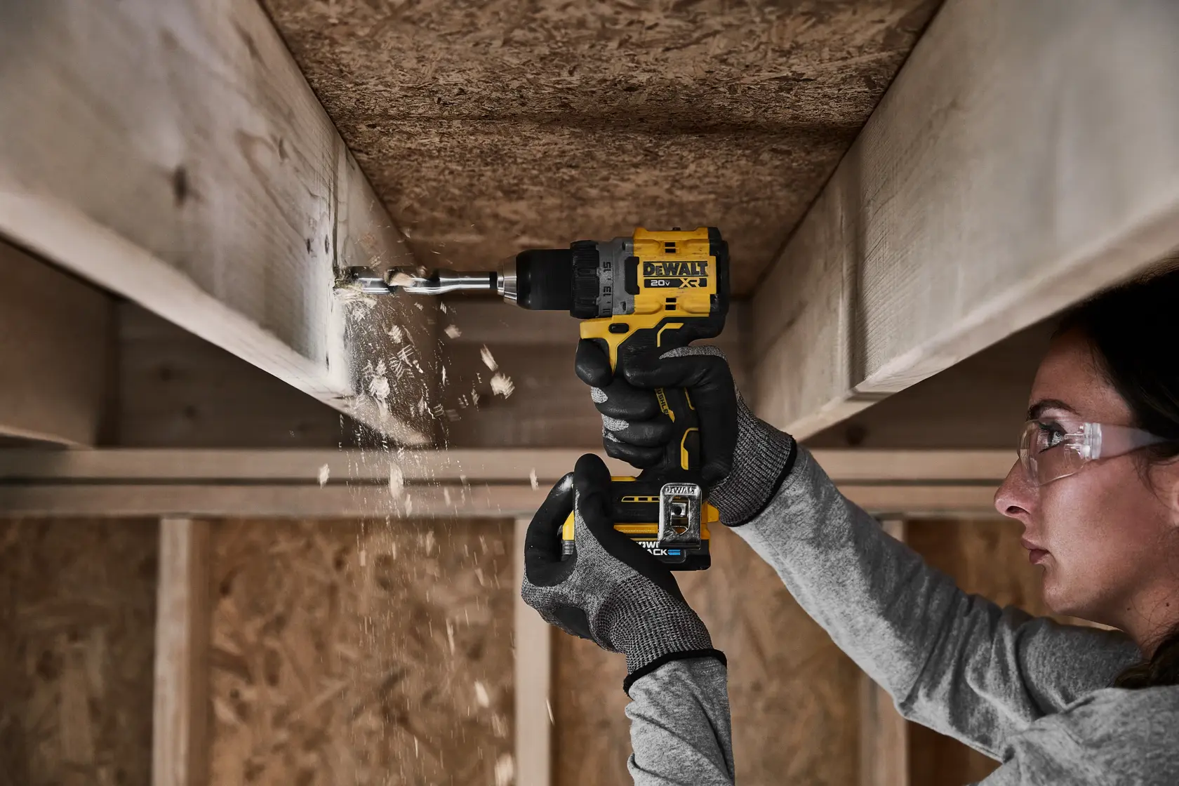Una persona con guantes está usando un taladro inalámbrico DEWALT para perforar una viga de madera, con virutas de madera cayendo visiblemente.