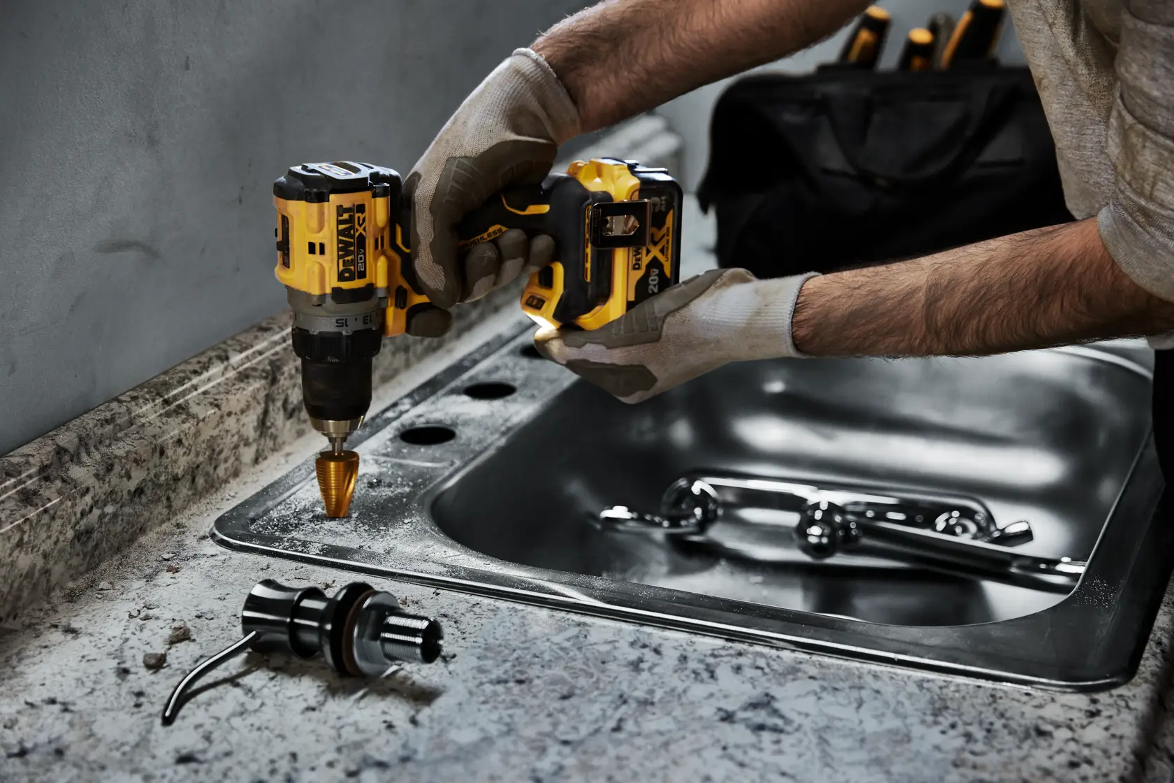 Una persona con guantes utiliza un taladro inalámbrico amarillo y negro DEWALT para perforar agujeros en una encimera cerca de un fregadero de acero inoxidable. Se puede ver una llave de agua y una pieza de fontanería en el mostrador. Una bolsa de herramientas negra está en el fondo.