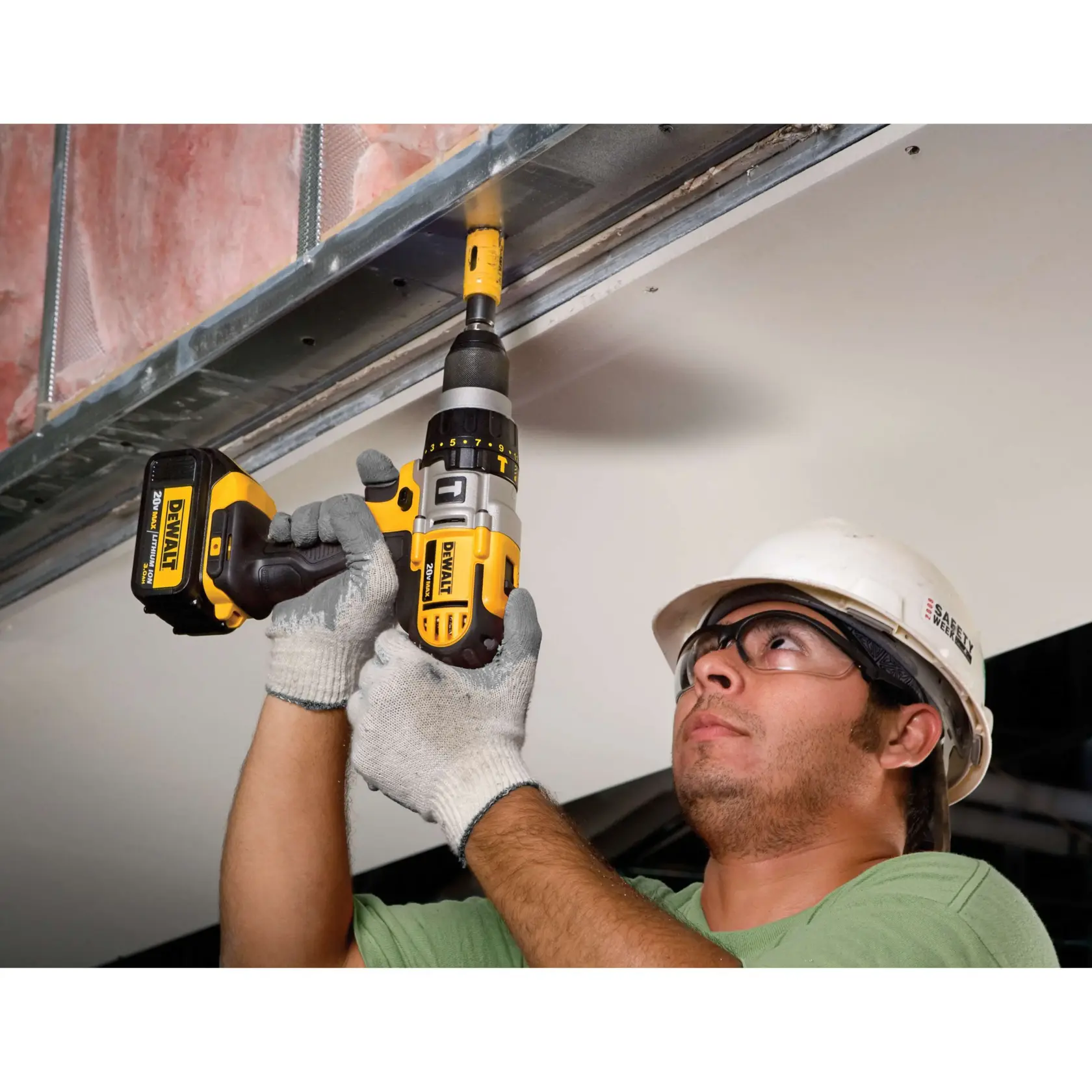 Una persona con guantes de seguridad y casco utiliza un taladro inalámbrico DEWALT DCD985L2 para instalar un tornillo en un marco de metal sobre un techo de placas de yeso.