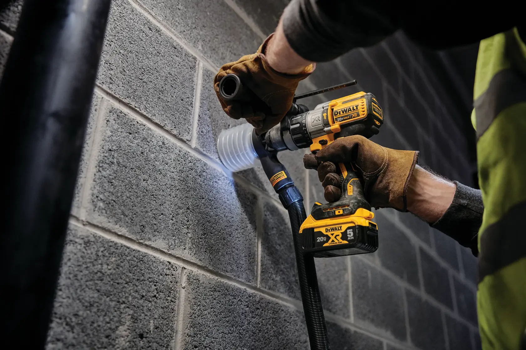 Una persona con guantes de trabajo marrones utiliza un taladro inalámbrico DEWALT amarillo y negro para perforar un agujero en una pared de bloques de hormigón gris. Hay un accesorio de extracción de polvo conectado al taladro.