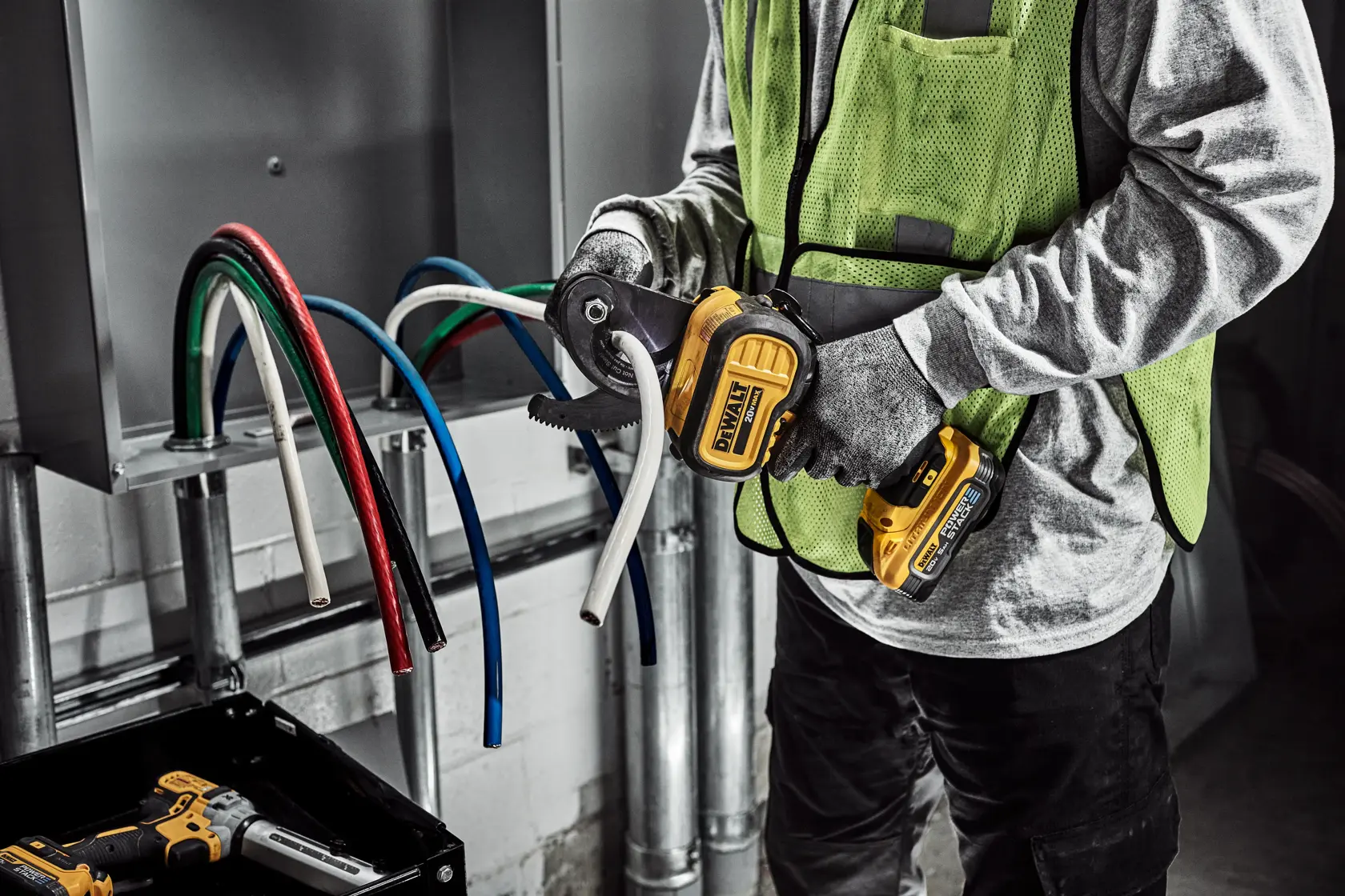 Una persona con guantes y chaleco de seguridad utiliza un cortador de cables inalámbrico DEWALT para cortar un cable eléctrico blanco grueso. Se pueden ver varios cables de colores saliendo de un panel eléctrico.