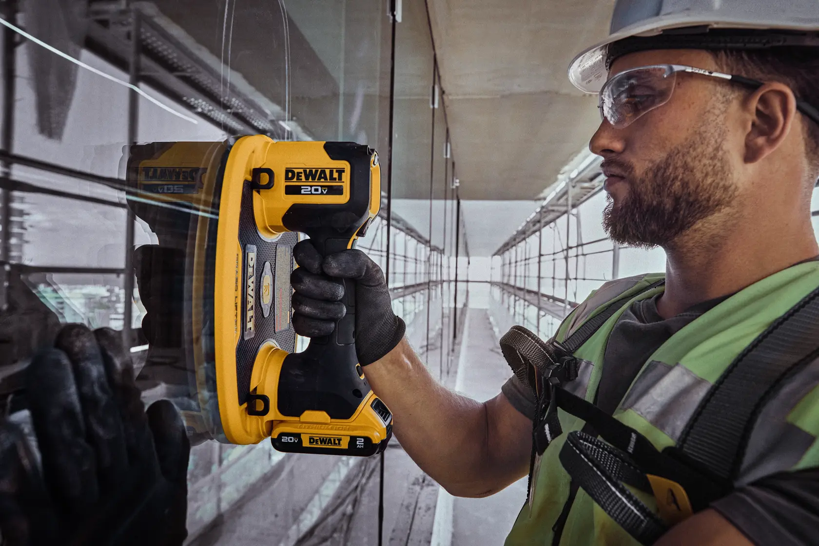 Una persona con equipo de seguridad está usando una herramienta portátil DEWALT 20V sobre una superficie de vidrio en un sitio de construcción, con el rostro difuminado por privacidad.