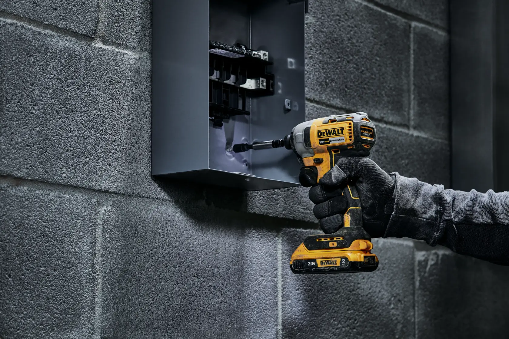 Un destornillador de impacto inalámbrico DEWALT usado por una mano enguantada para trabajar en un panel eléctrico montado en una pared de concreto.