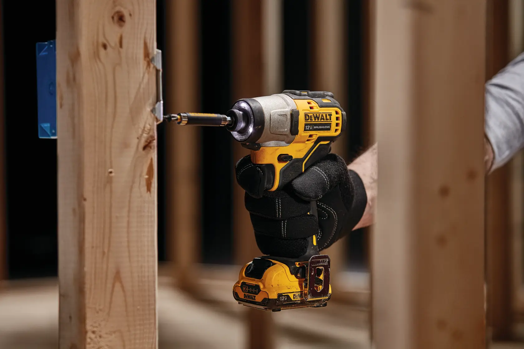 Una persona con guantes negros utiliza un destornillador de impacto inalámbrico amarillo DEWALT para atornillar un tornillo en una estructura de madera durante una construcción.