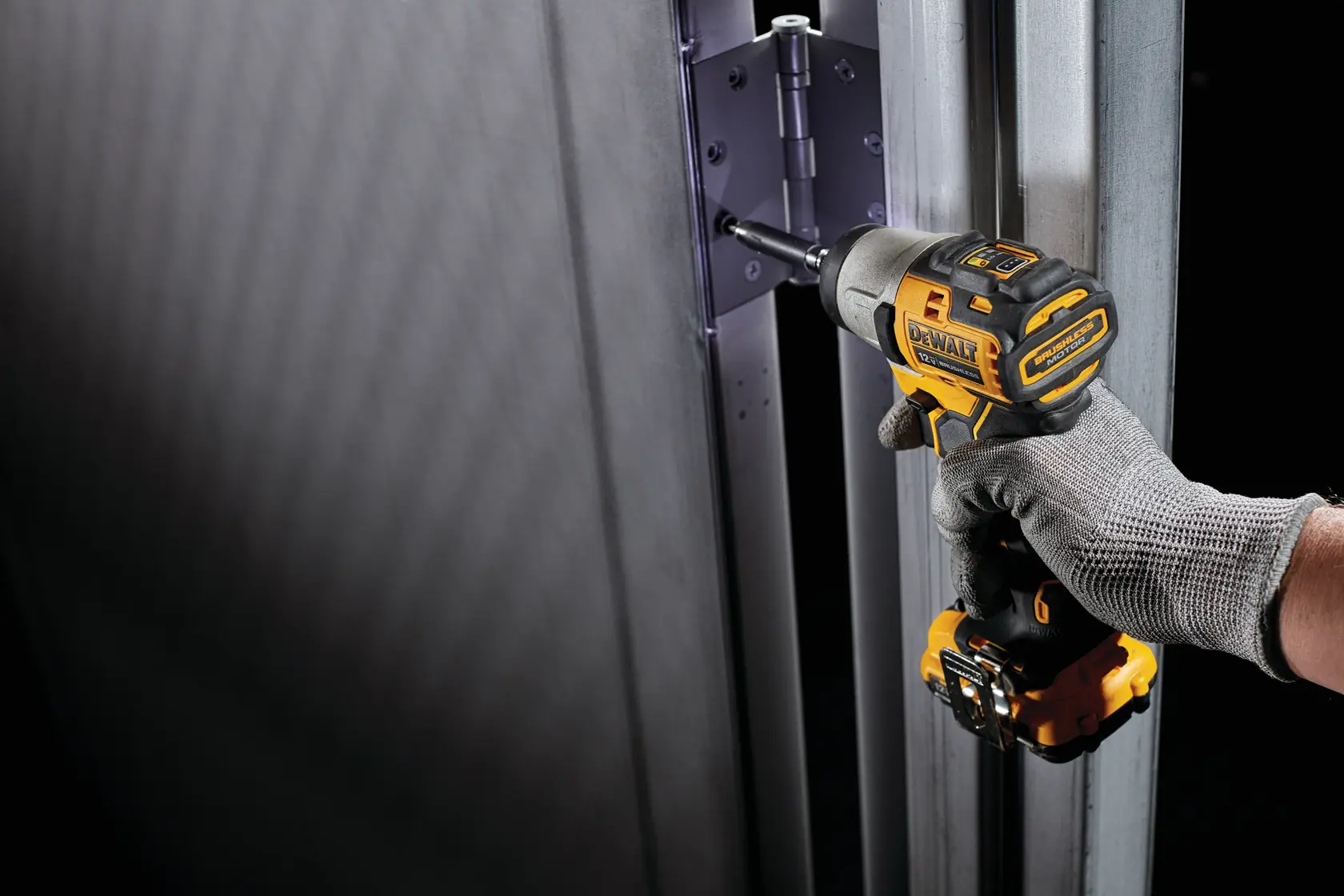 Una persona usando un guante de trabajo gris utiliza un destornillador de impacto inalámbrico DEWALT para atornillar una bisagra en un marco de puerta de metal.