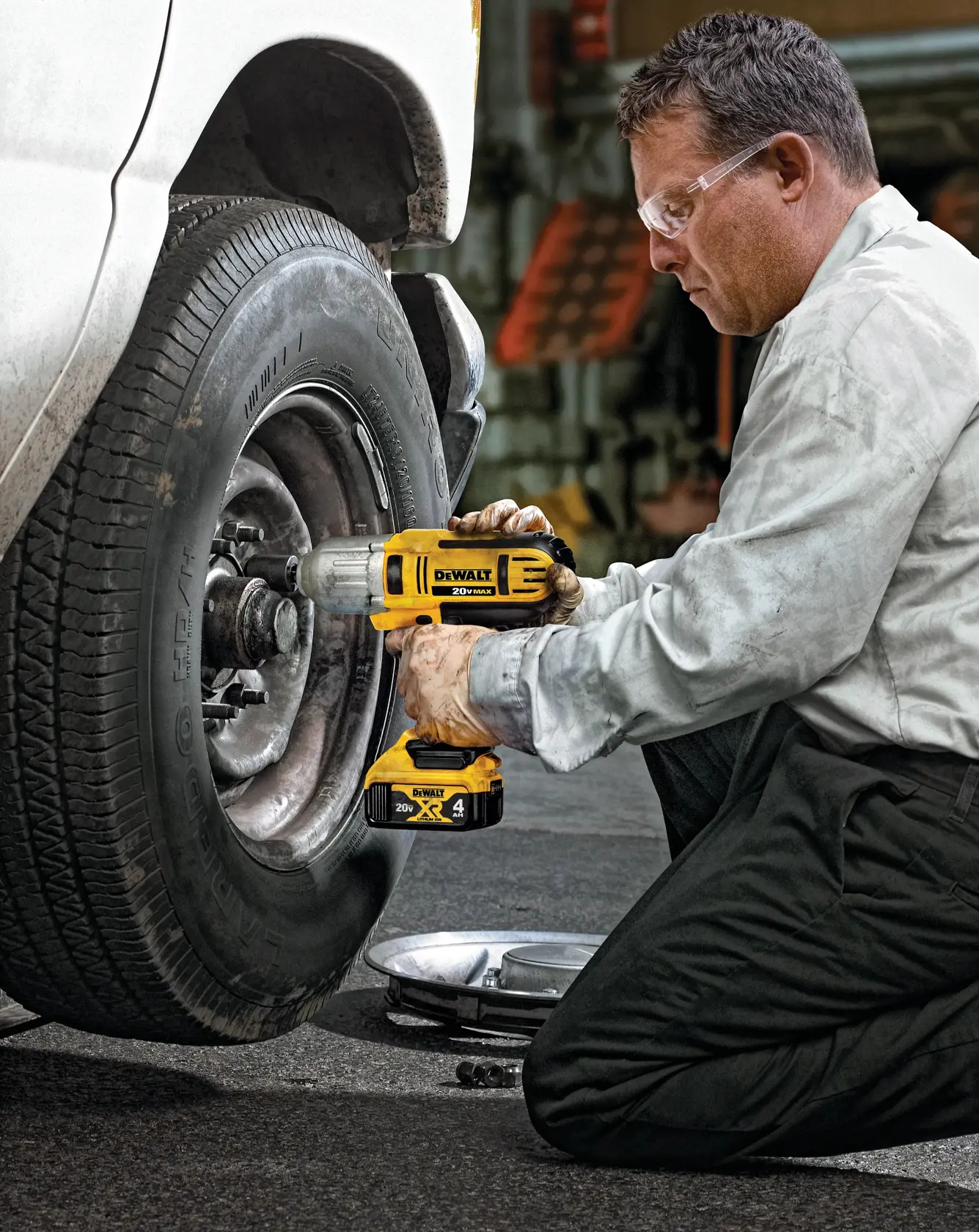 Una persona con el rostro desenfocado está arrodillada y usando una llave de impacto inalámbrica DEWALT para aflojar o apretar las tuercas de una rueda de coche. El vehículo está estacionado y la escena parece ser en un taller o garaje.