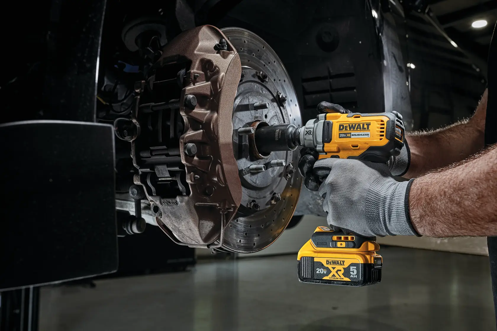 Una persona usa una llave de impacto inalámbrica DEWALT 20V XR para aflojar o apretar tuercas de rueda en un conjunto de disco de freno de vehículo. La herramienta es amarilla y negra y la persona lleva guantes de trabajo.