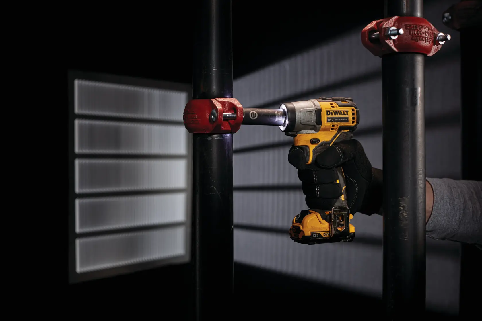 Una persona con un guante negro está usando un destornillador de impacto inalámbrico DEWALT para apretar un tornillo en un accesorio de tubería metálico. La herramienta es amarilla y negra, y parece ser utilizada en un entorno industrial o de construcción.