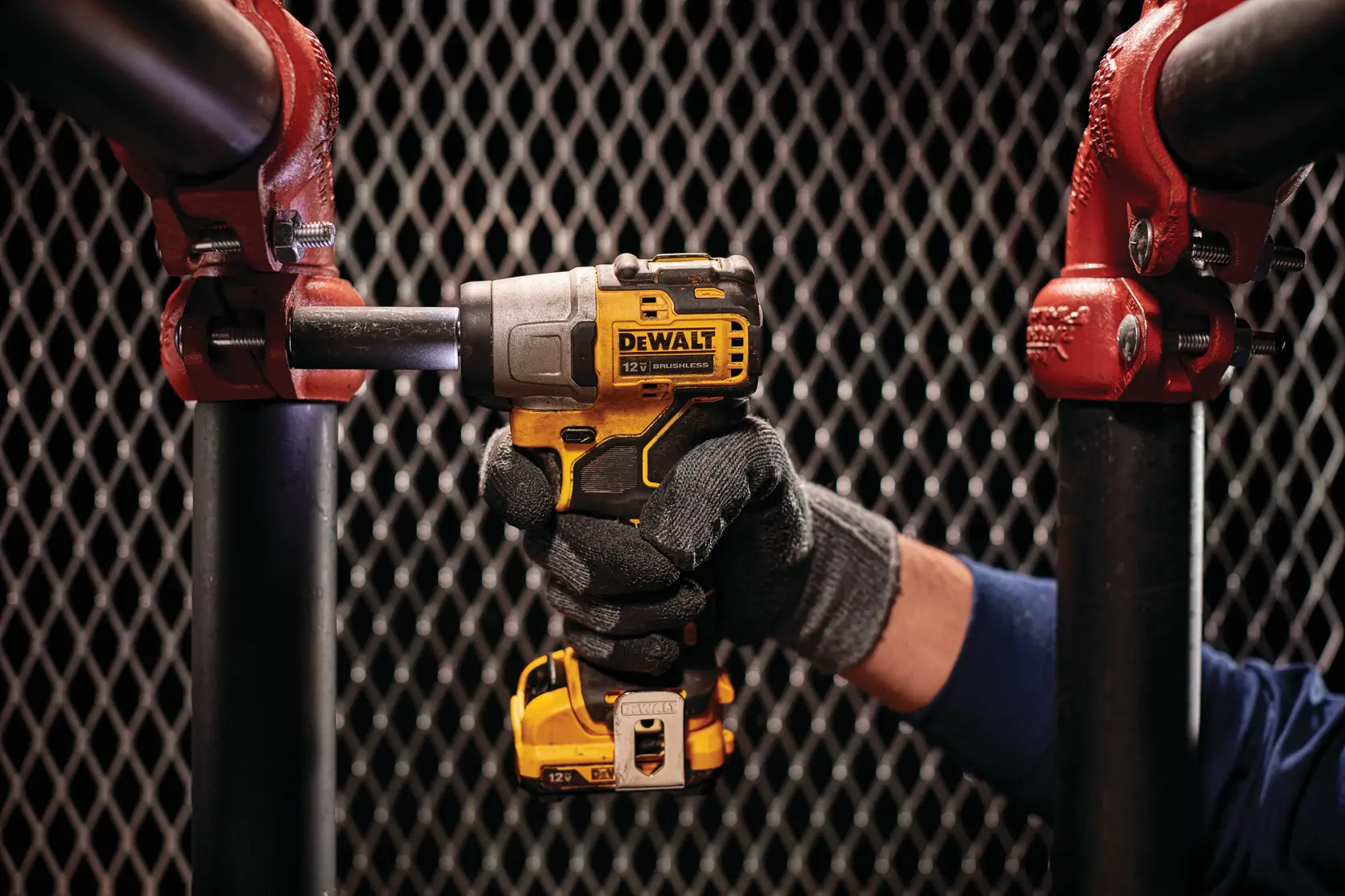 Una persona con guantes utiliza una llave de impacto inalámbrica DEWALT de 12V en una gran unión de tuberías de metal negro, con un fondo de malla.