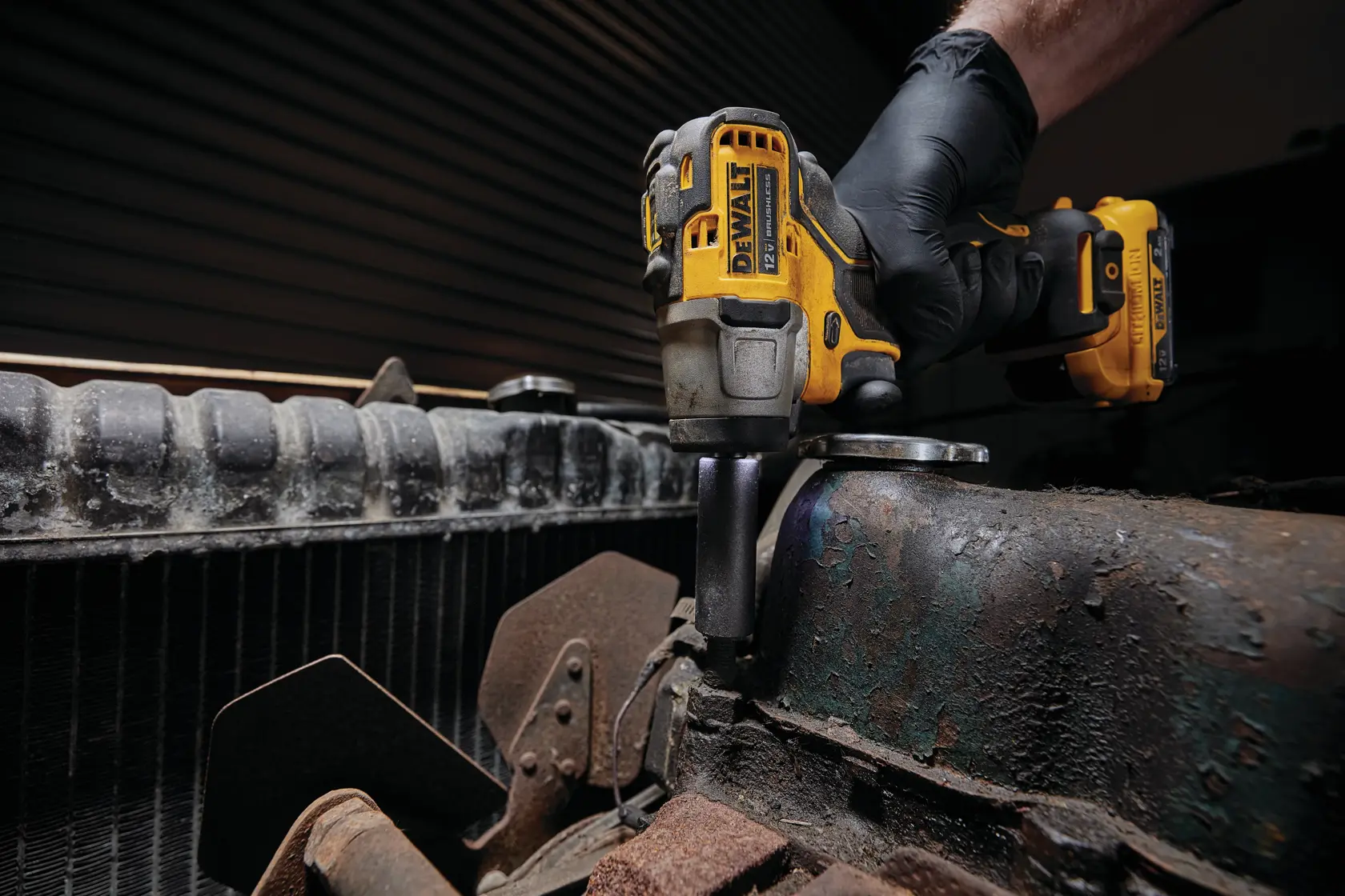 Una persona con un guante negro usa una llave de impacto inalámbrica DEWALT DCF902B para trabajar en un componente de motor oxidado.