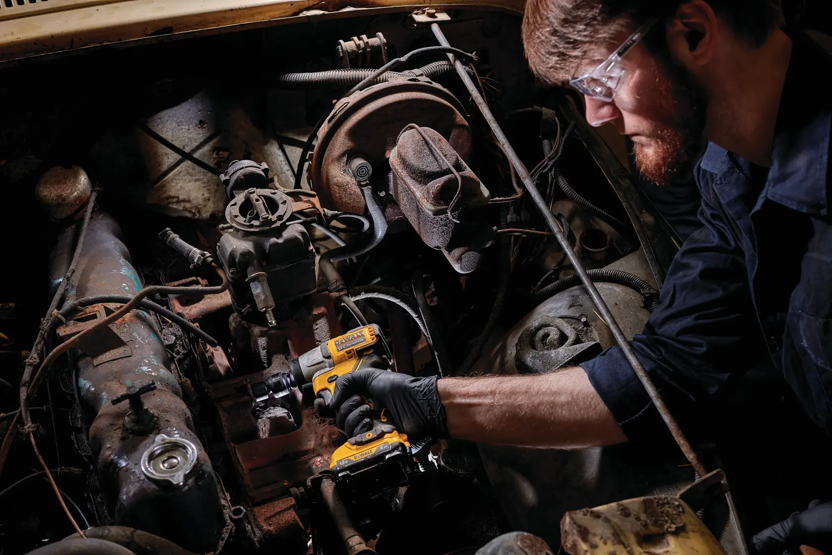Una persona con guantes está usando una llave de impacto inalámbrica DEWALT DCF902B para trabajar en el motor de un vehículo. El rostro de la persona está oculto y el enfoque está en la herramienta y los componentes del motor.