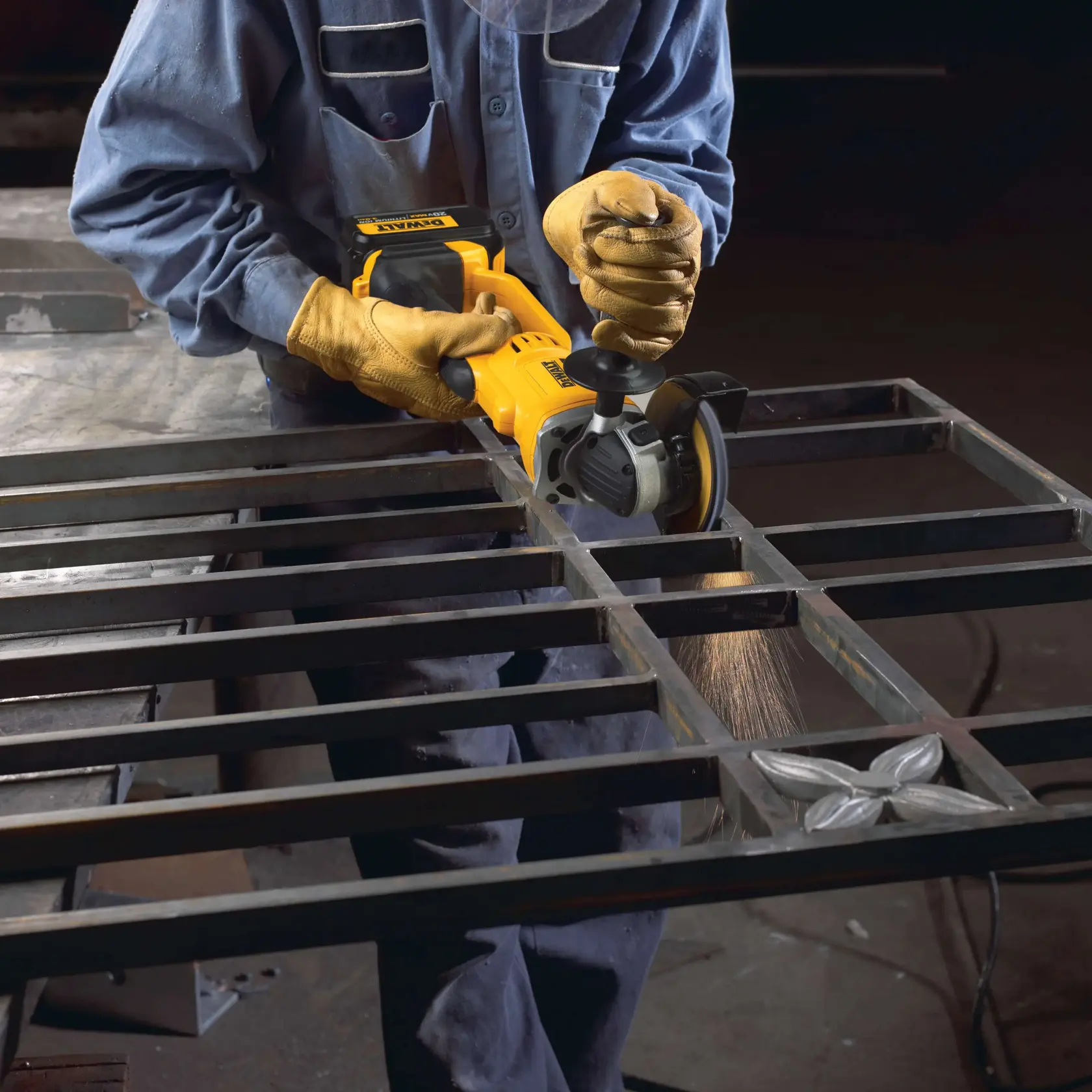 Una persona con guantes protectores y un uniforme de trabajo azul está utilizando una amoladora angular inalámbrica DEWALT (modelo DCG412B) para lijar o cortar una estructura de rejilla metálica. Se ven chispas en el punto de contacto entre la herramienta y el metal.