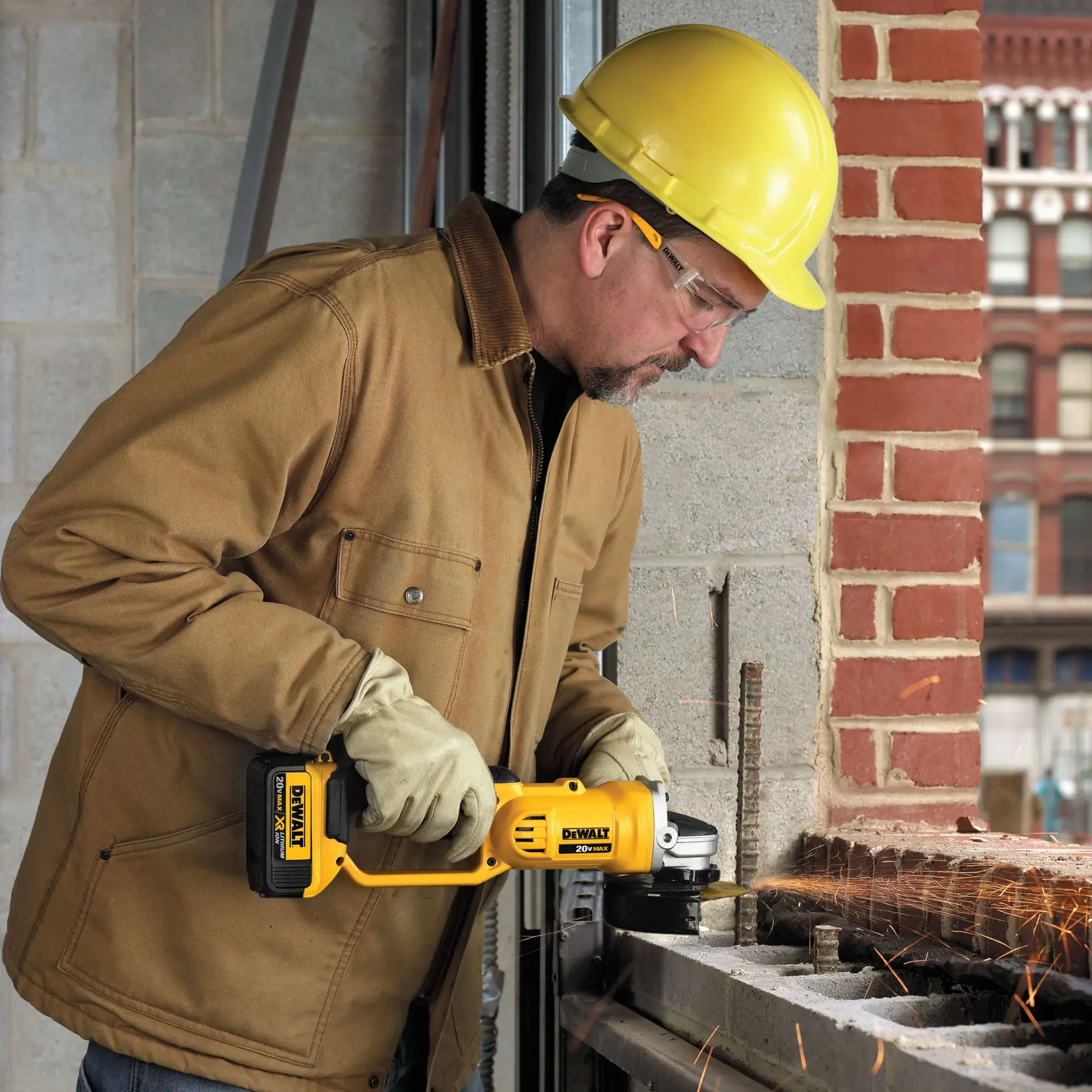 Una persona que lleva chaqueta de trabajo marrón, guantes y casco amarillo está usando una amoladora inalámbrica DEWALT (modelo DCG412M2) para cortar metal en un sitio de construcción, con chispas volando desde la zona de corte.
