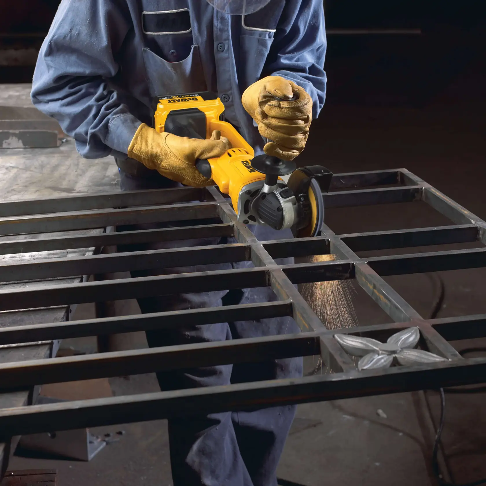 Una persona con guantes protectores y un uniforme de trabajo azul utiliza una amoladora angular amarilla DEWALT para suavizar o cortar el metal sobre un marco metálico soldado, mientras saltan chispas.